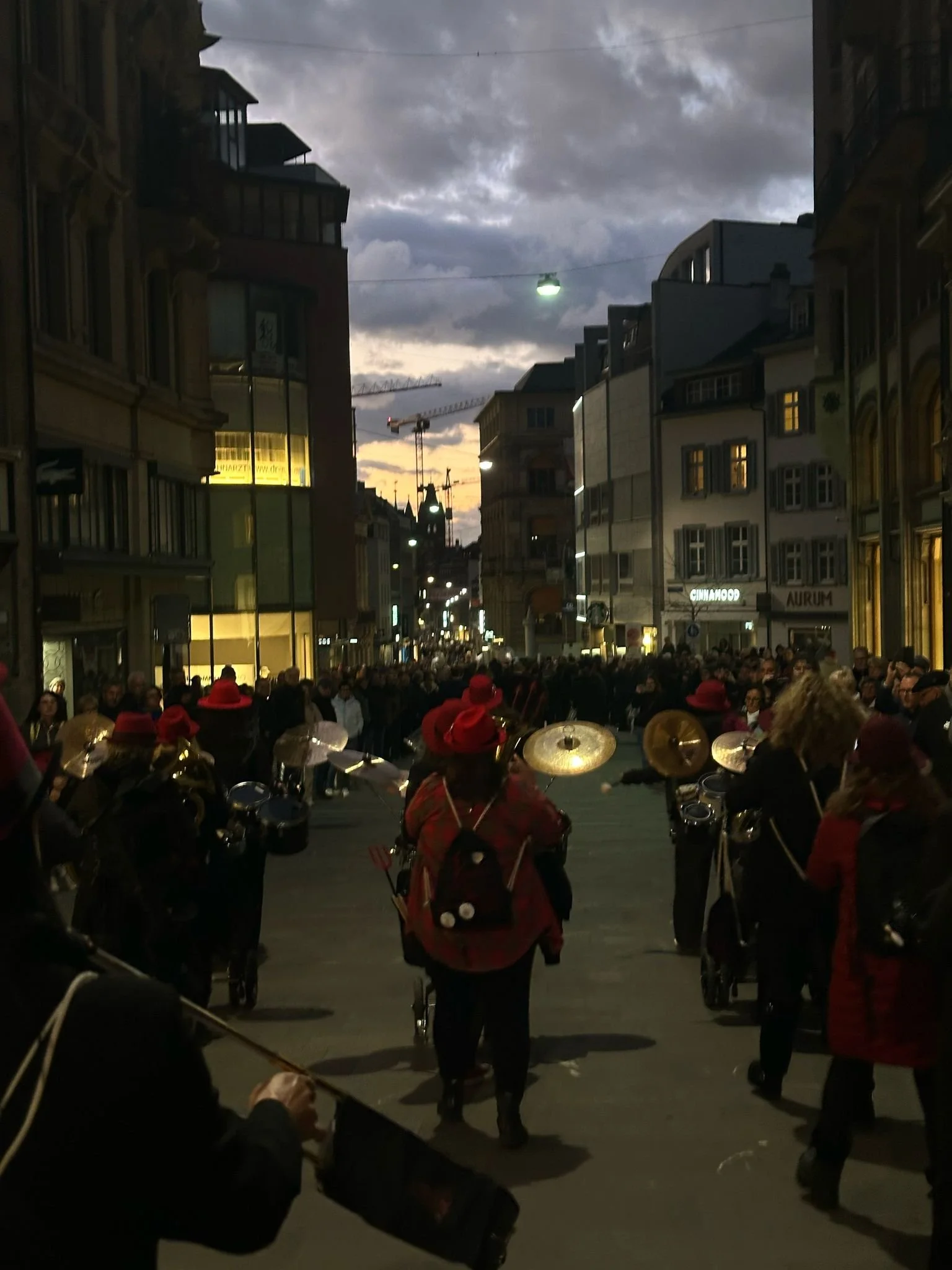 Straßenszene in einer Stadt bei Dämmerung mit einer Parade von Musikern, die Trommeln spielen, Menschen in roten Hüten, Mehrfamilienhäuser und Wolken am Himmel.