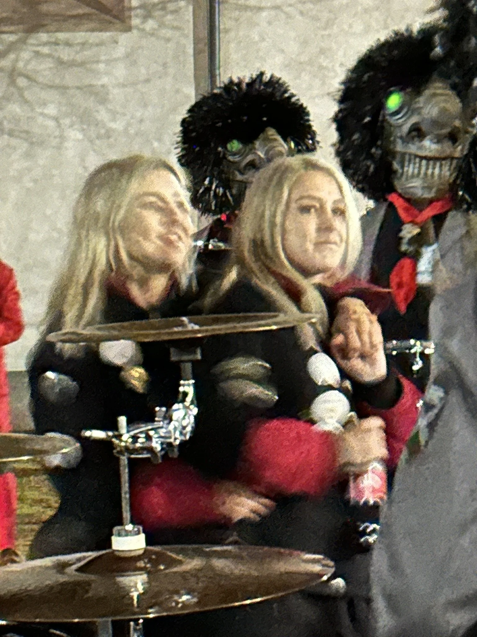 Menschen in Kostümen mit Masken und Perücken, eine Person hält einen Regenschirm, sie sitzen hinter einem Schlagzeug.