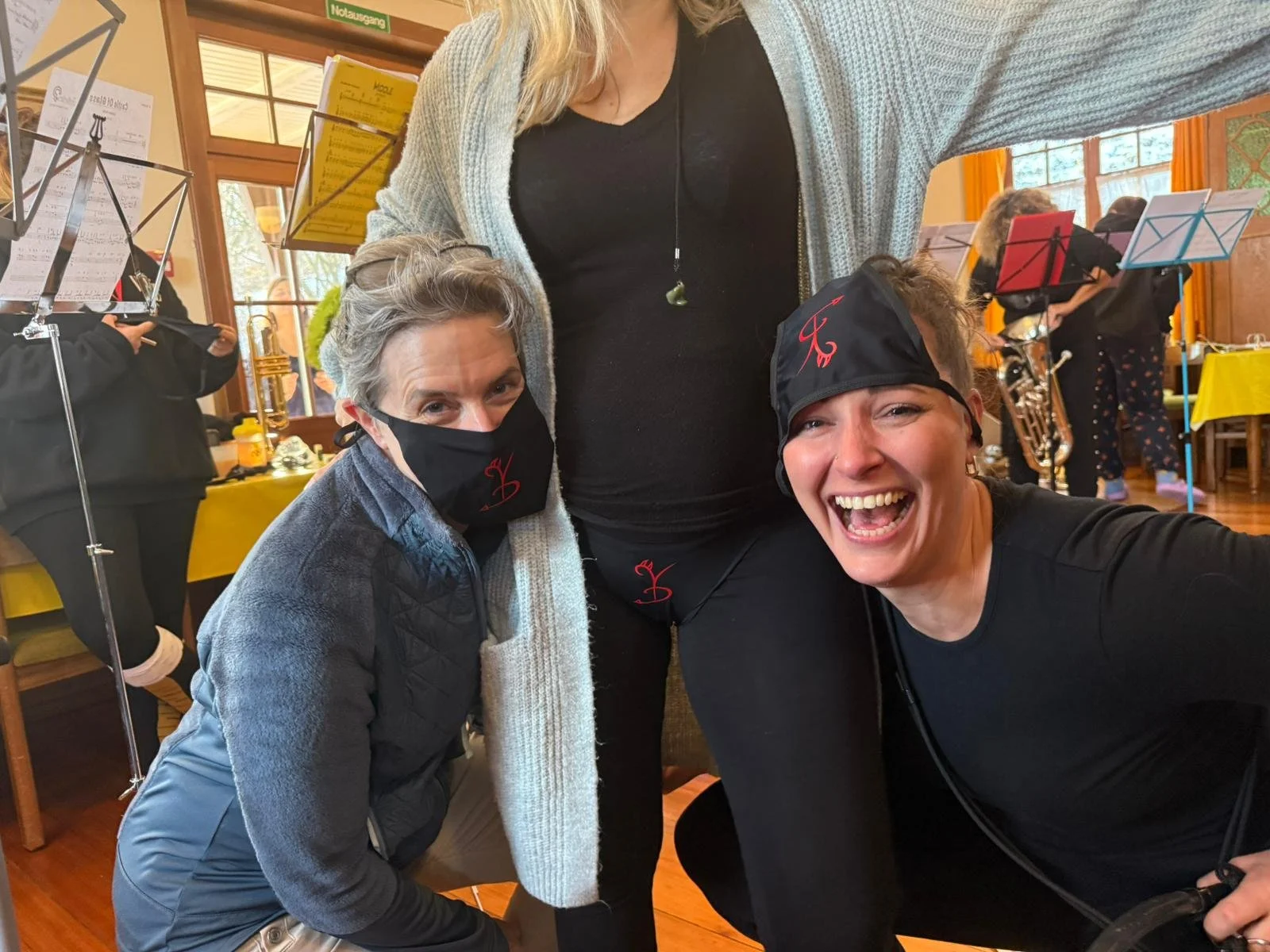 Drei lachende Frauen in einem Raum, eine trägt eine Maske, die anderen beiden sind ohne Maske, im Hintergrund Musiker mit Musikständern und Instrumenten, gelbe Tischtücher auf Tischen.