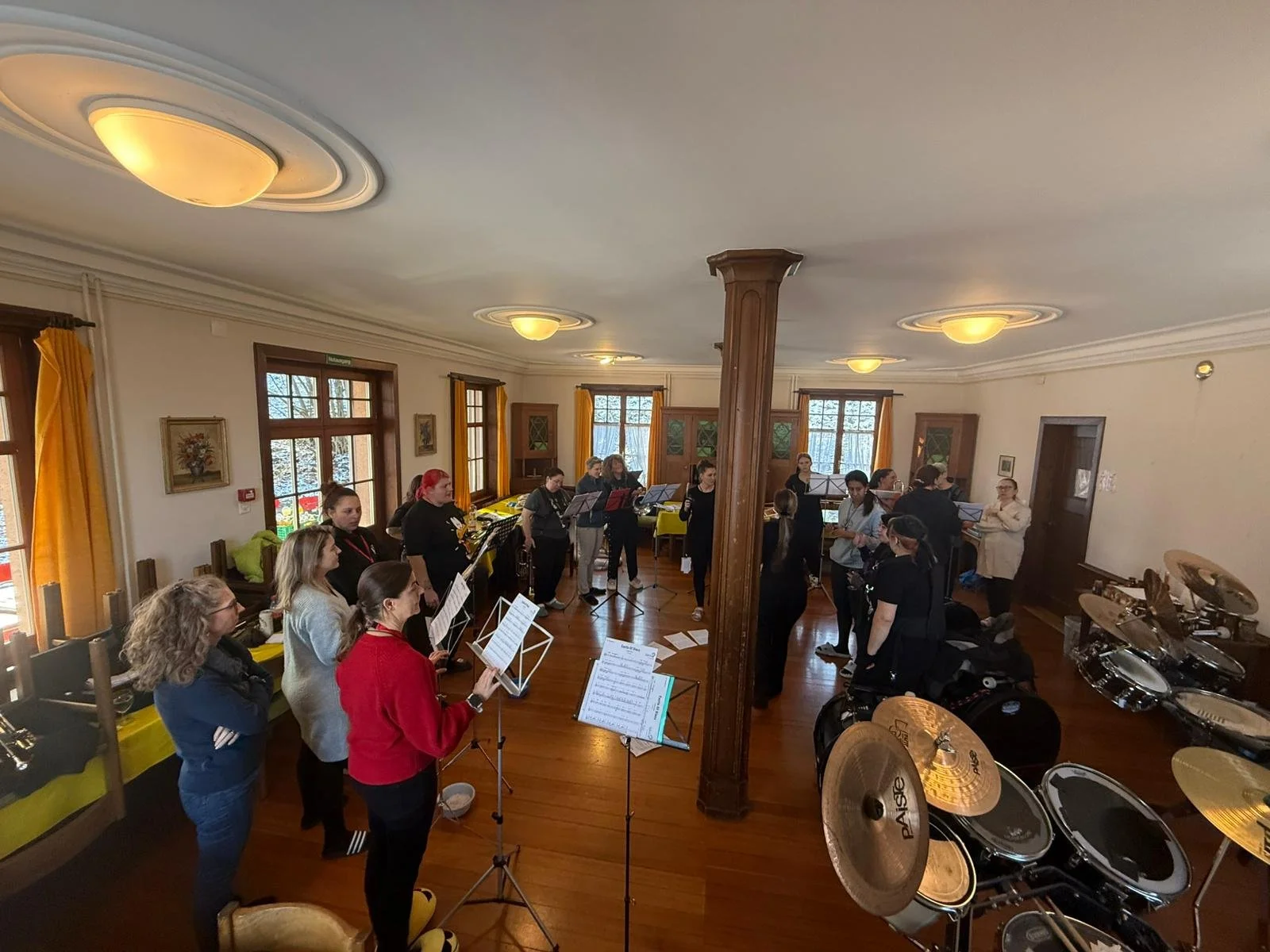 Musiker in einem Raum mit Holzfußboden, spielen Musik während eines Proben- oder Orchesterunterrichts, einige mit Notenblättern, an der Wand hängen Bilder, im Raum stehen Trommeln und andere Musikinstrumente.