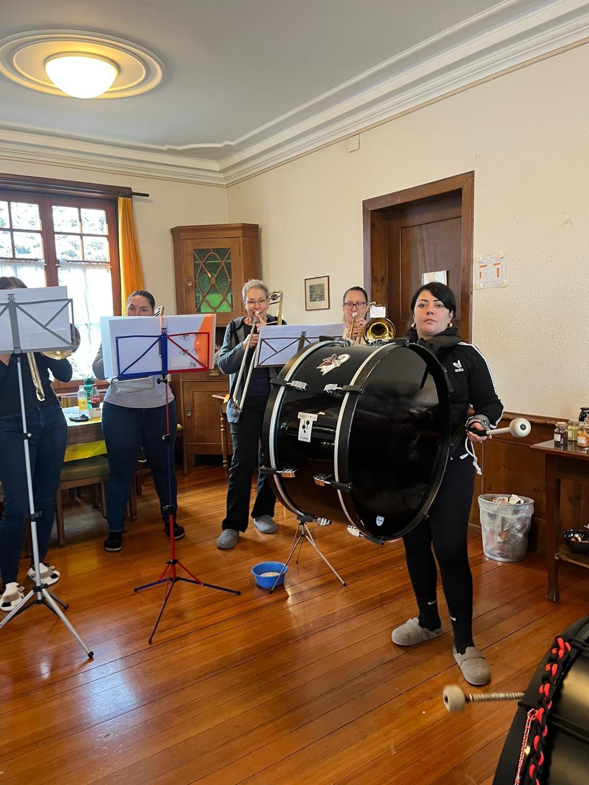 Musikerinnen mit Blasinstrumenten und einer großen Trommel in einem gemütlichen Raum mit Holzfußboden, Fenster und Holzschrank.