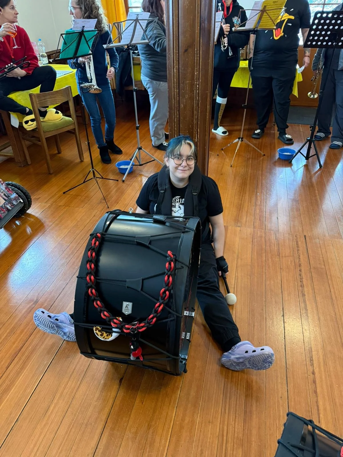 Ein Mädchen sitzt im Boden, umgeben von einer großen schwarzen Kiste, die mit einer roten und schwarzen Kordel befestigt ist. Sie trägt eine schwarze Jacke, eine schwarze Hose, Turnschuhe und Brille, und hält einen Schläger mit einem weißen Ball. Im 