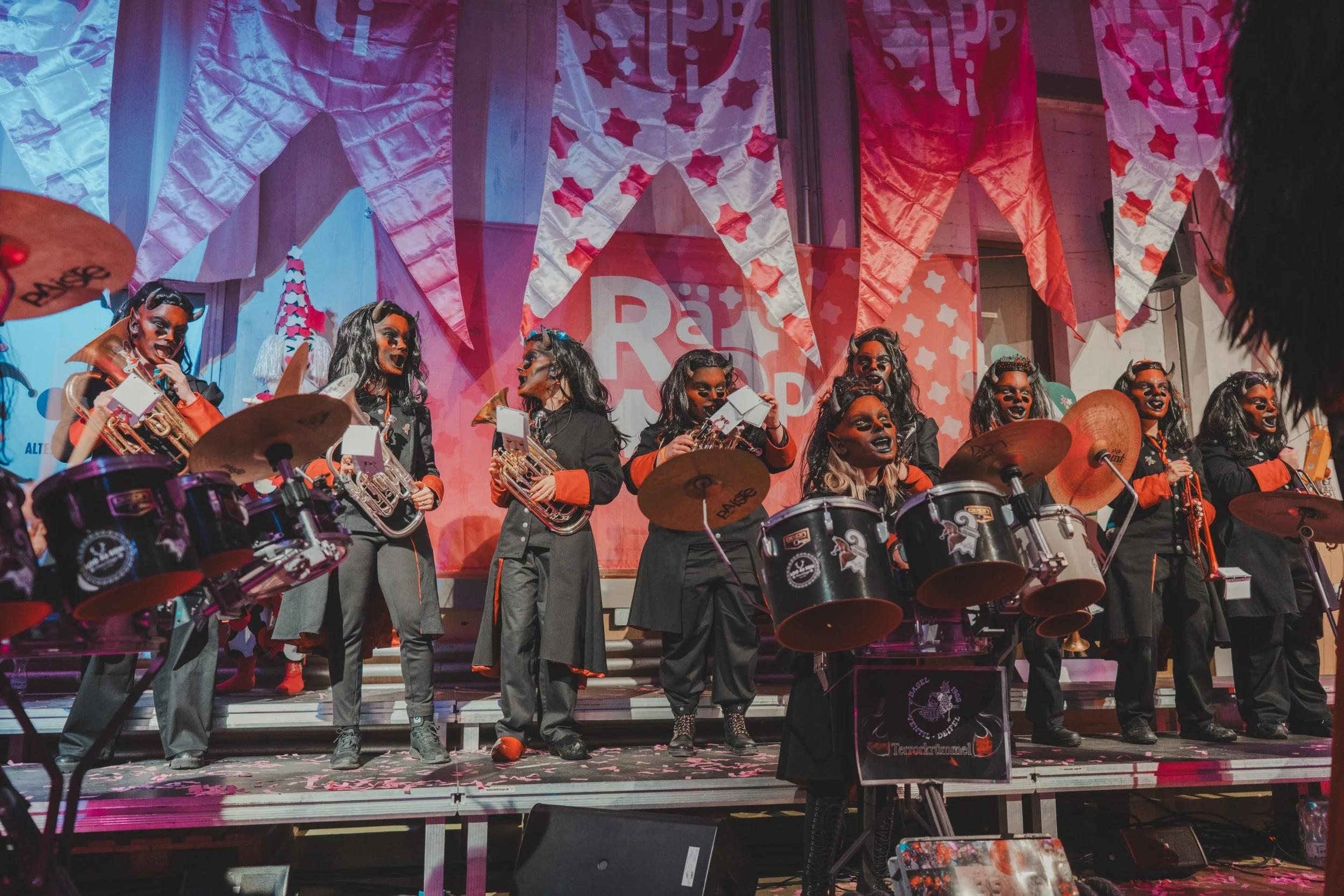 Eine Gruppe von Musikern auf einer Bühne, die in Auftrittskostümen mit schwarzen und orangenen Outfits, schwarzem Make-up und Hörnern auf dem Kopf ein Brass- oder Blasorchester spielt.