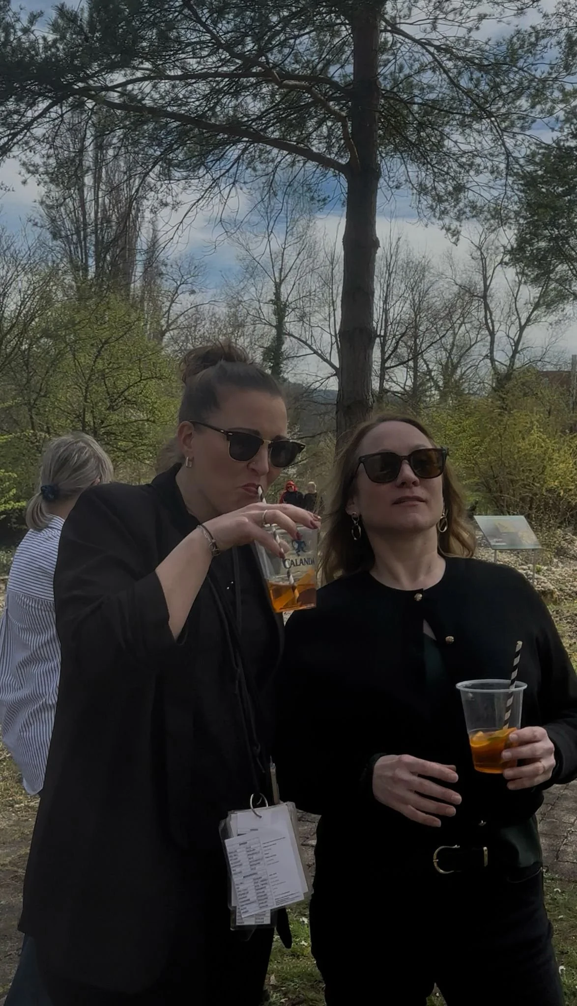 Zwei Frauen mit dunklen Sonnenbrillen trinken im Freien Getränke, im Hintergrund Bäume und ein bewölkter Himmel.