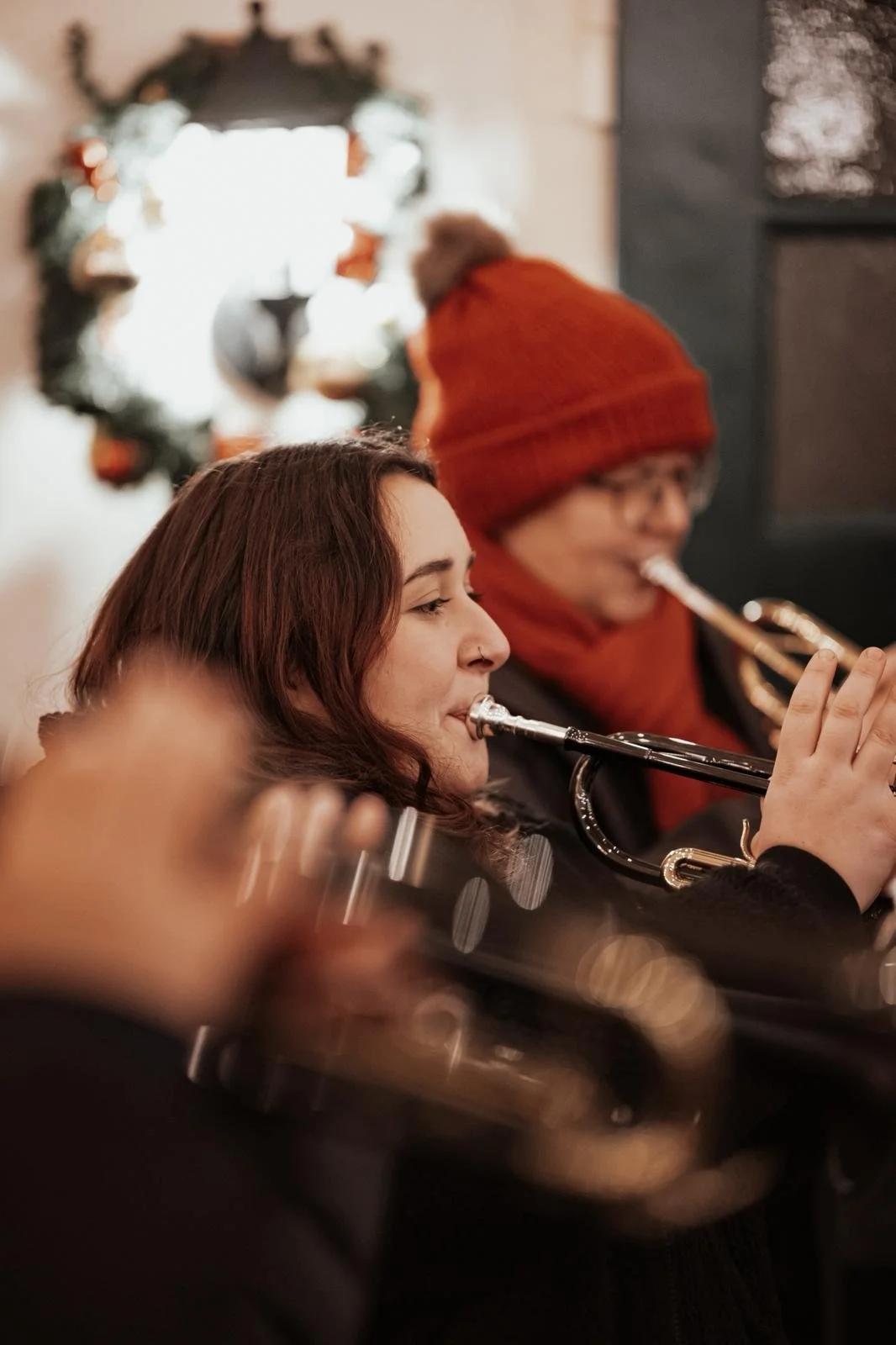 Menschen spielen im Winter Weihnachtslieder mit Trompeten, eine Frau mit langen braunen Haaren, eine Person mit roter Mütze und Brille, im Hintergrund Weihnachtsdekoration.