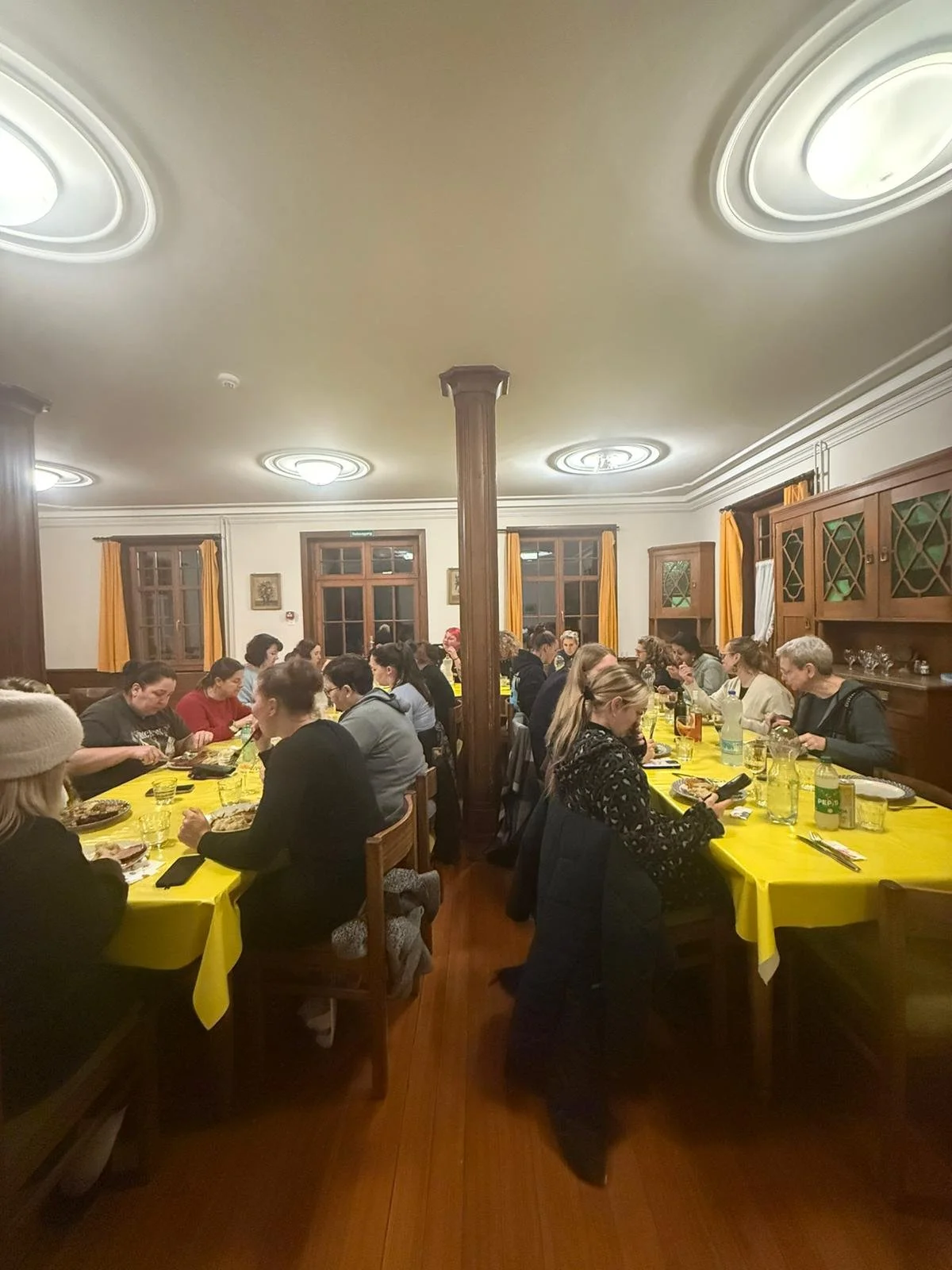 Mehrere Menschen sitzen an zwei langen Tischen mit gelben Tischtüchern und essen in einem gut beleuchteten Raum mit Holzdecke, Fenstern mit gelben Vorhängen und Holzregalen.