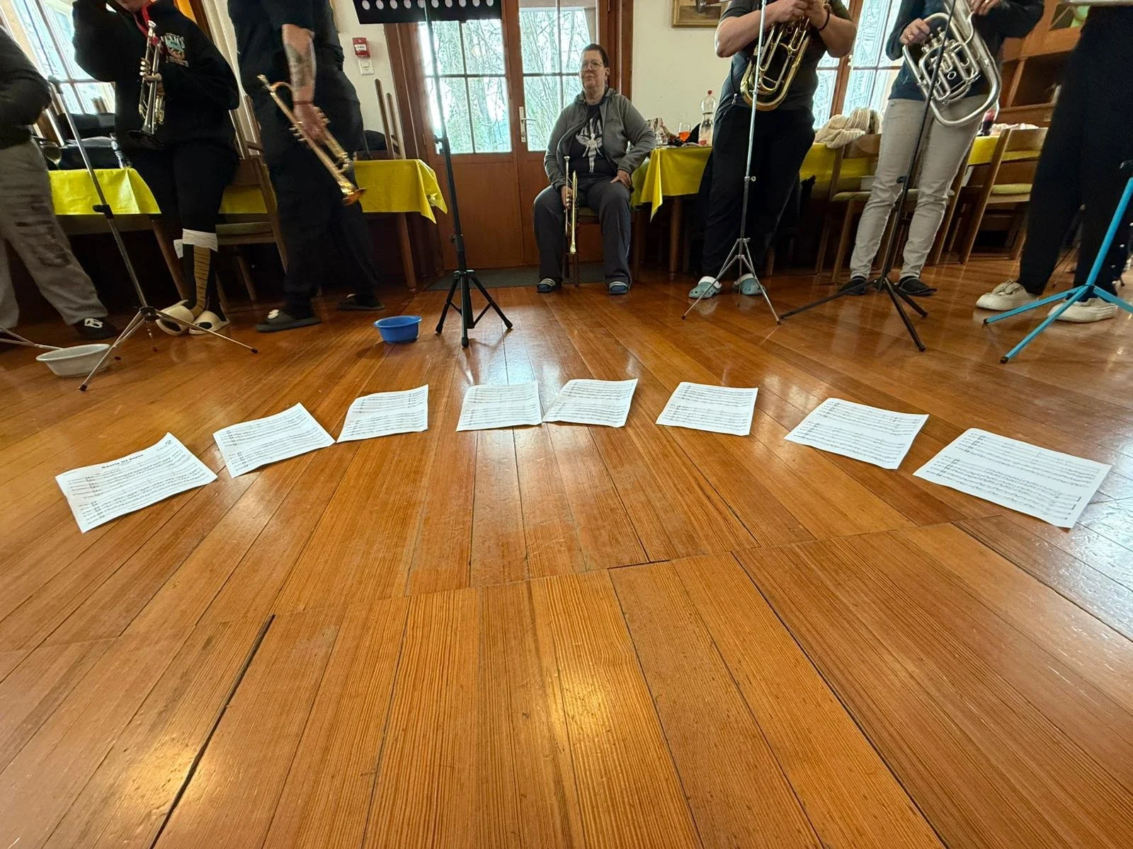 Musiker proben in einem Raum mit Holzboden, auf dem verstreute Notenblätter liegen, während sie ihre Instrumente spielen und Musik notieren.
