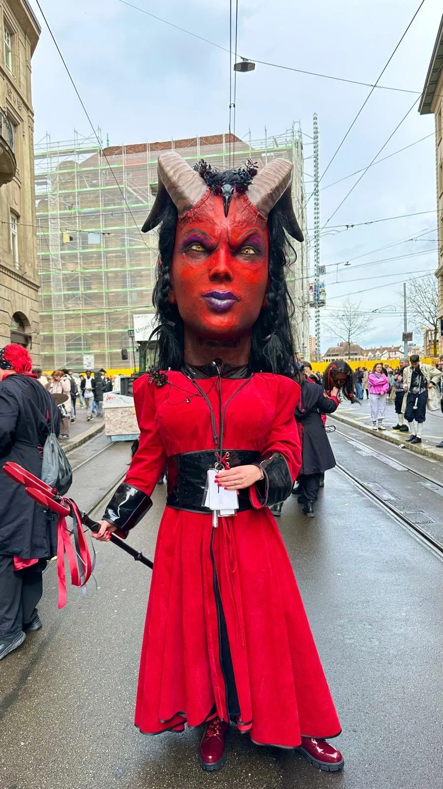 Eine Person in Karnevals- oder Faschenkostüm mit Dämonengesicht, Hörnern und rotem Kleid auf einer Straße während eines Festivals.