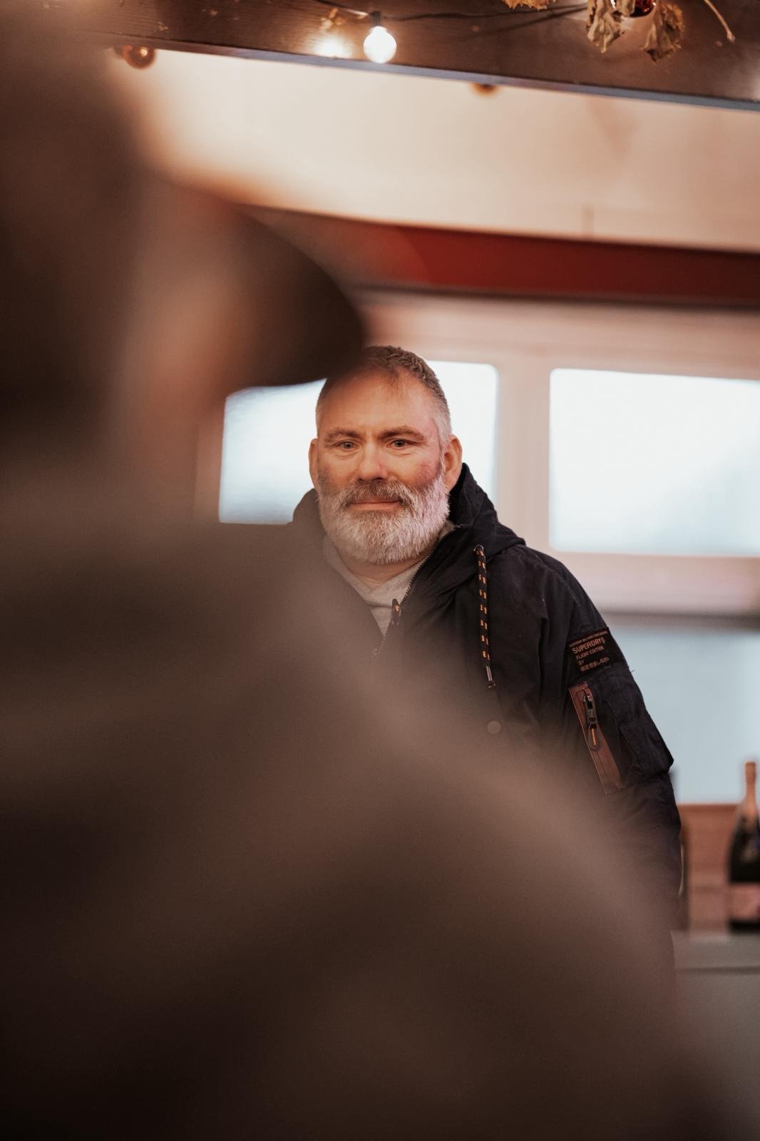 Ein Mann mit grauem Bart und Kurzhaar schaut in die Kamera, im Hintergrund zwei Fenster, im Vordergrund unscharfe Objekte, möglicherweise eine Person oder Gegenstände.