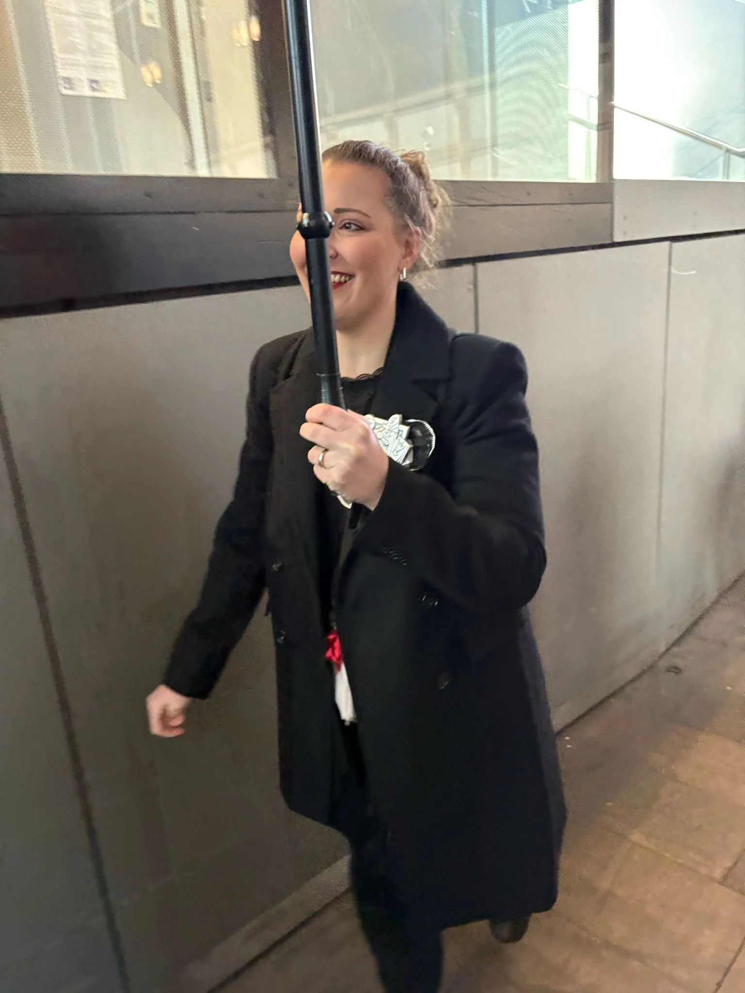 Eine lachende Frau mit einem schwarzen Mantel, die einen Regenschirm hält, geht an einer grauen Wand entlang in einem städtischen Umfeld.