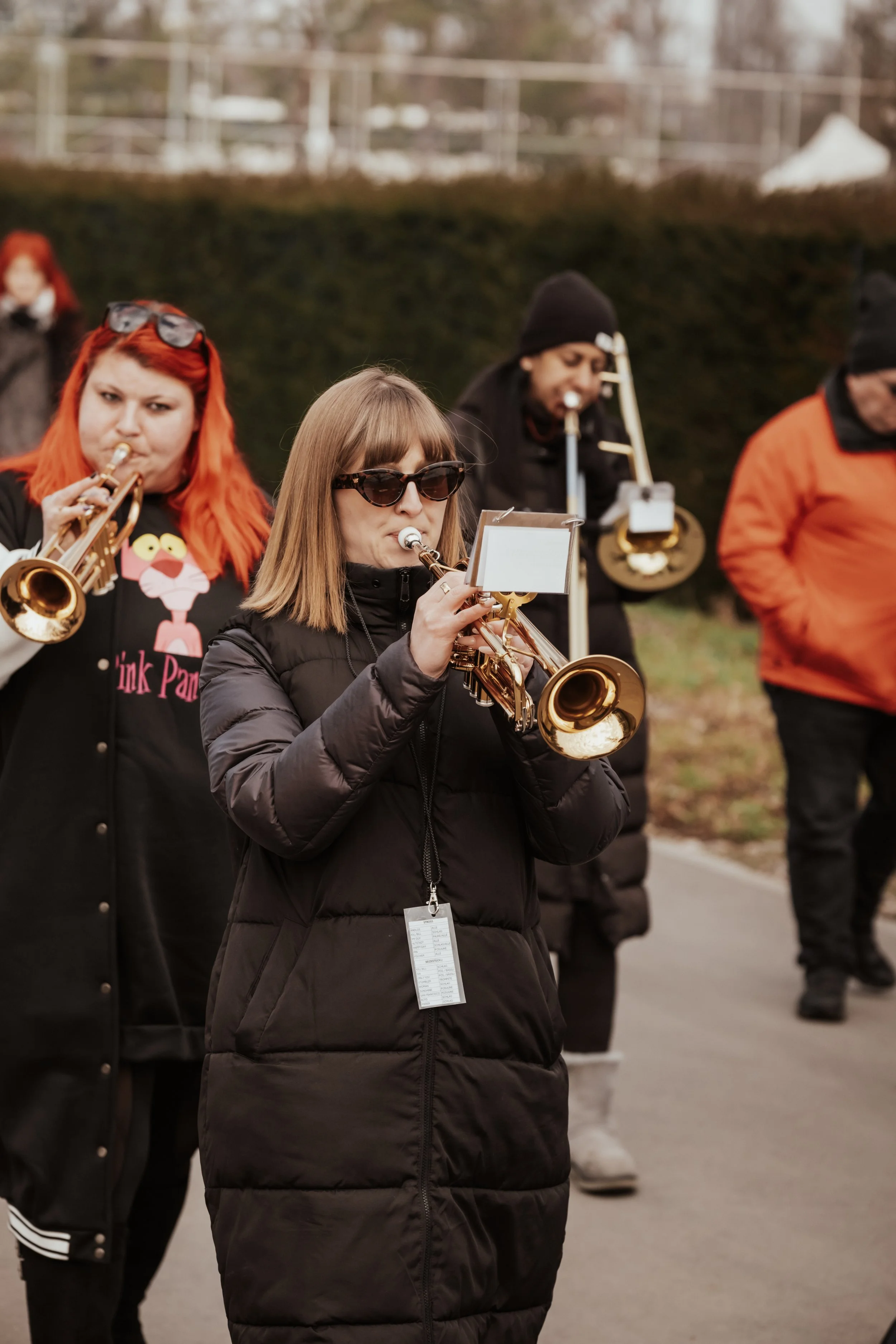 Frauen spielen Trompeten und eine Posaune bei einem Außenauftritt, eine trägt Sonnenbrille, andere Musiker sind im Hintergrund, alle tragen Winterkleidung.