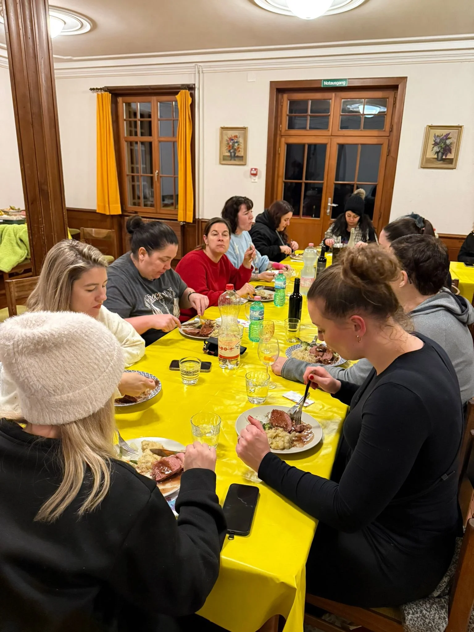 Mehrere Menschen sitzen an einem langen Tisch mit gelber Tischdecke und essen. Es sind etwa zehn Frauen unterschiedlichen Alters, einige trinken, andere essen Fleischgerichte, auf dem Tisch stehen Flaschen Wasser und Wein. Es scheint sich um eine Diä