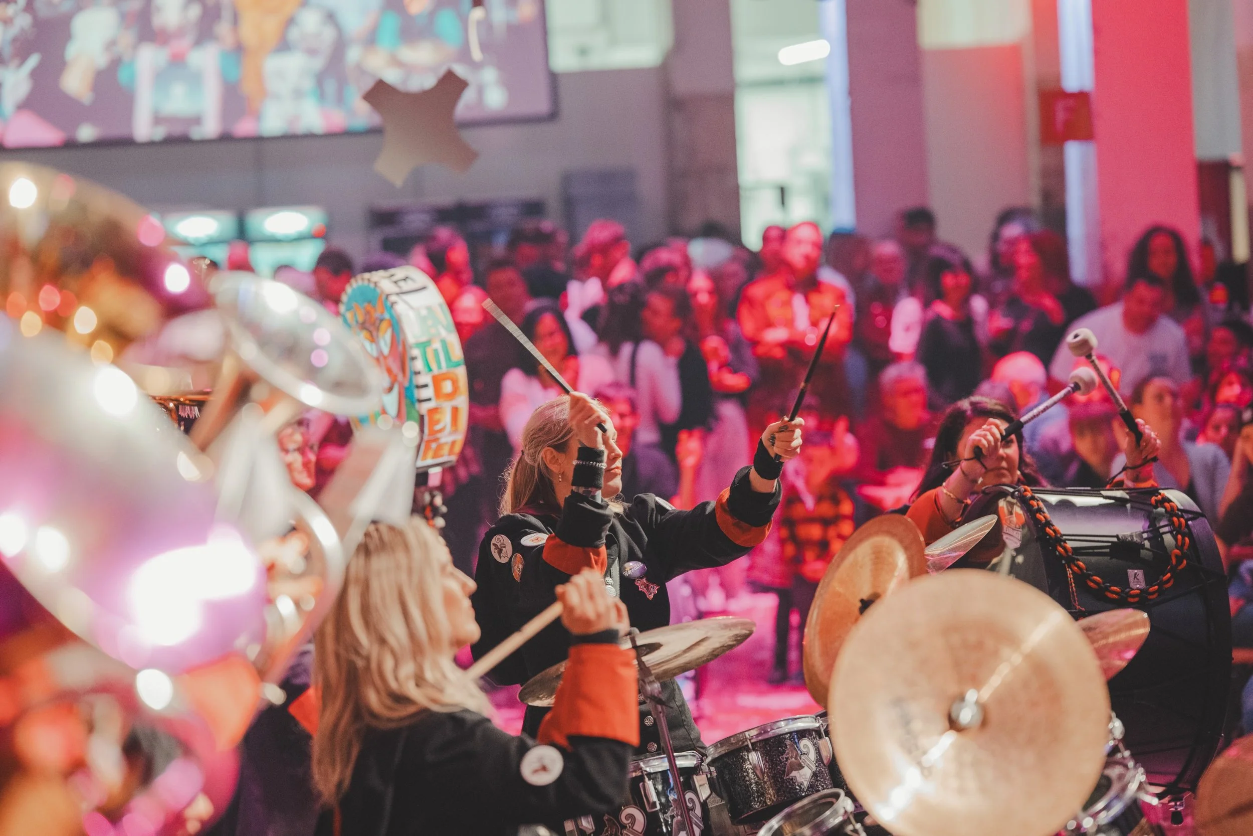 Musikerinnen spielen Schlagzeug bei einer Veranstaltung mit einer großen Zuschauermenge im Hintergrund, festliche Dekorationen und helle Lichter.