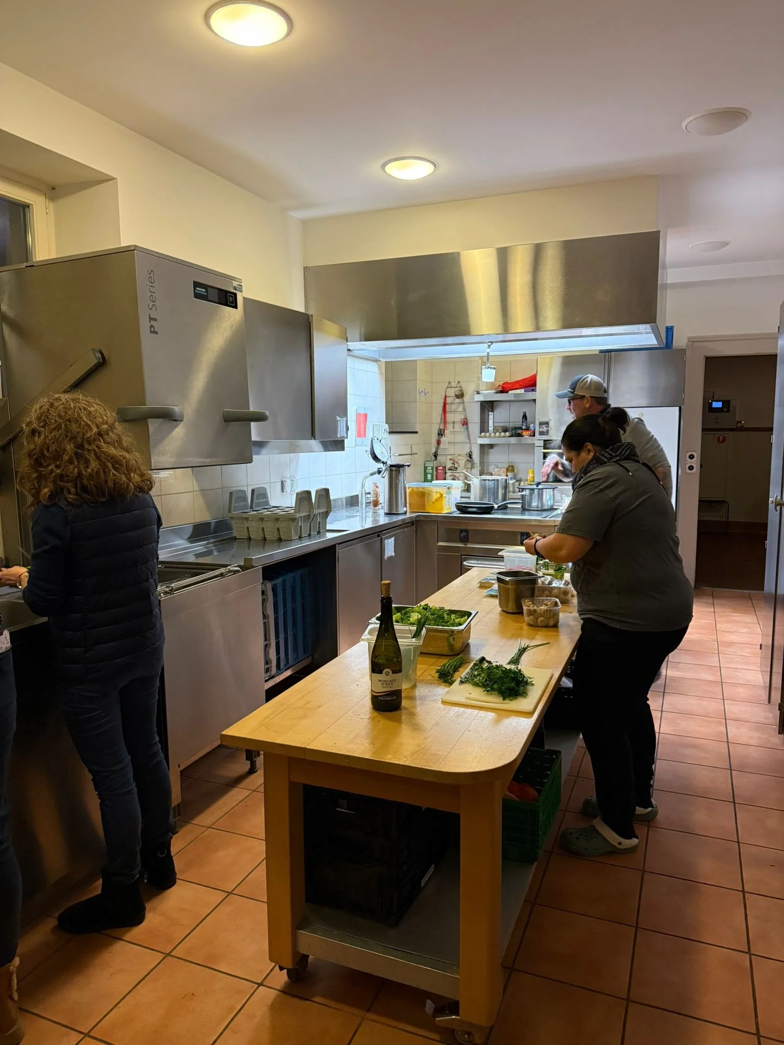 Menschen arbeiten in einer professionellen Küche mit Holzarbeitsplatte, auf der frische Kräuter liegen, während sie verschiedene Kochvorgänge ausführen.