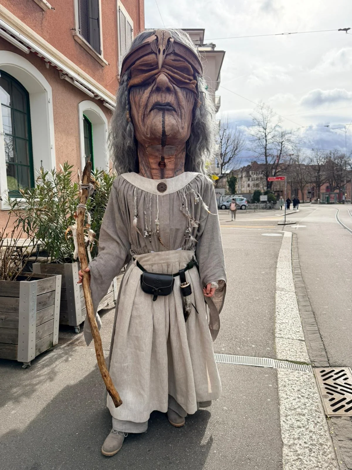Eine Person trägt eine große Maske oder ein Kostüm mit einem alten, hölzernen Gesicht und einer Krone auf dem Kopf. Sie trägt ein langes, beige Gewand und hält einen Stock. Die Person steht auf einer Straße in einer Stadt.