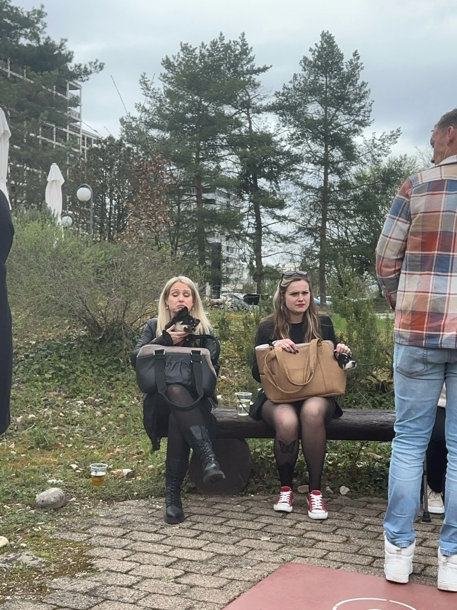 Zwei junge Frauen sitzen auf einer Bank im Freien, eine trägt schwarze Kleidung und einen schwarzen Hund, die andere trägt eine schwarze Jacke und hat eine braune Tasche. Zwei Männer sind in der Nähe, einer trägt einen karierten Hemd. Es gibt Bäume, 