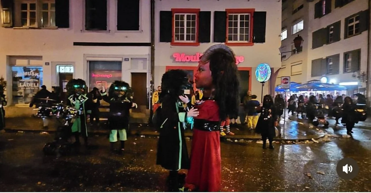 Zwei große Figuren, wahrscheinlich Maskottchen, stehen sich in der Mitte der Straße gegenüber. Im Hintergrund sind Menschen, die vermutlich bei einer Feier oder Parade teilnehmen, und Gebäude mit beleuchteten Fenstern und Schildern.