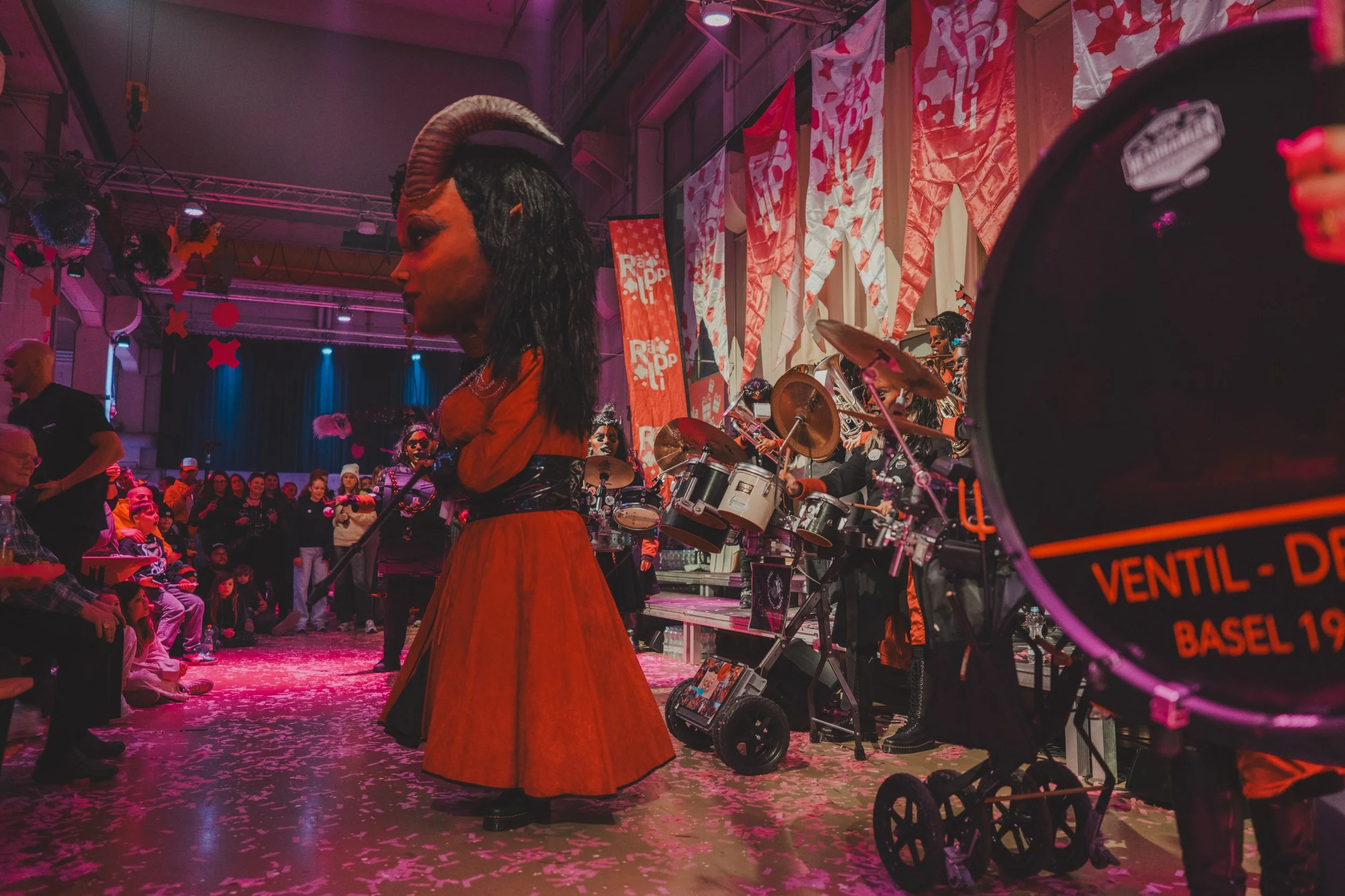 Karnevalsveranstaltung mit jemandem in einer großen Dekomasse nach Karneval, die mit großen, bunten Figuren, Trommeln und einer Horde von Menschen im Hintergrund aufstehen. Der Raum ist mit roten und weißen Flaggen und Bannern geschmückt, die den kar