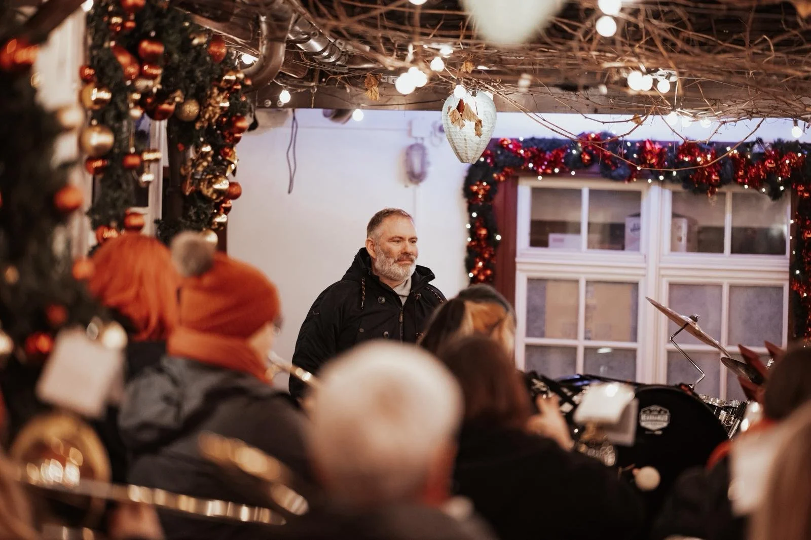 Menschen, die sich bei einer festlichen Weihnachtsfeier versammeln, dekoriert mit Weihnachtsgirlanden und Lichterketten, und einem Musiker, der hinter einem Schlagzeug sitzt, während die Fenster im Hintergrund erleuchtet sind.