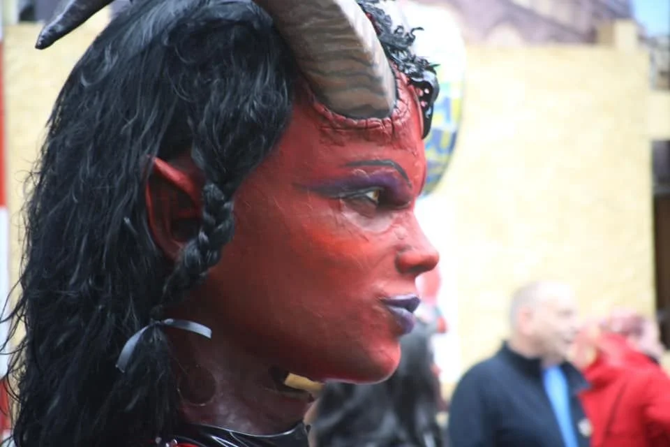 Person in Teufelskostüm mit roten Gesicht, Hörnern, schwarzen Haaren und gedrehtem Zopf, die in Profilansicht zu sehen ist, im Hintergrund Menschen in einem Raum.