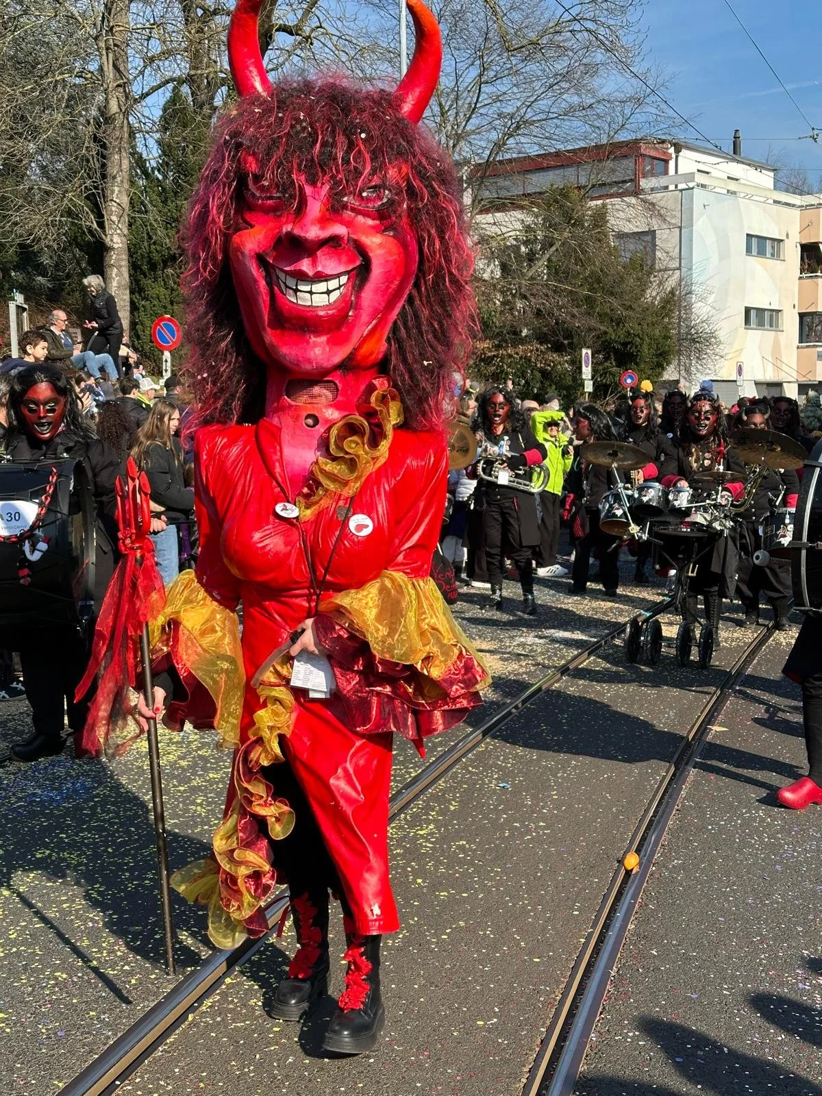 Person in teufelhaftigem Kostüm mit großen roten Hörnern, rotem Gesicht und braunem Lockenkopf läuft während eines Straßenkarnevals. Weitere Teilnehmer in ähnlichen Kostümen sind im Hintergrund mit Musikinstrumenten zu sehen.