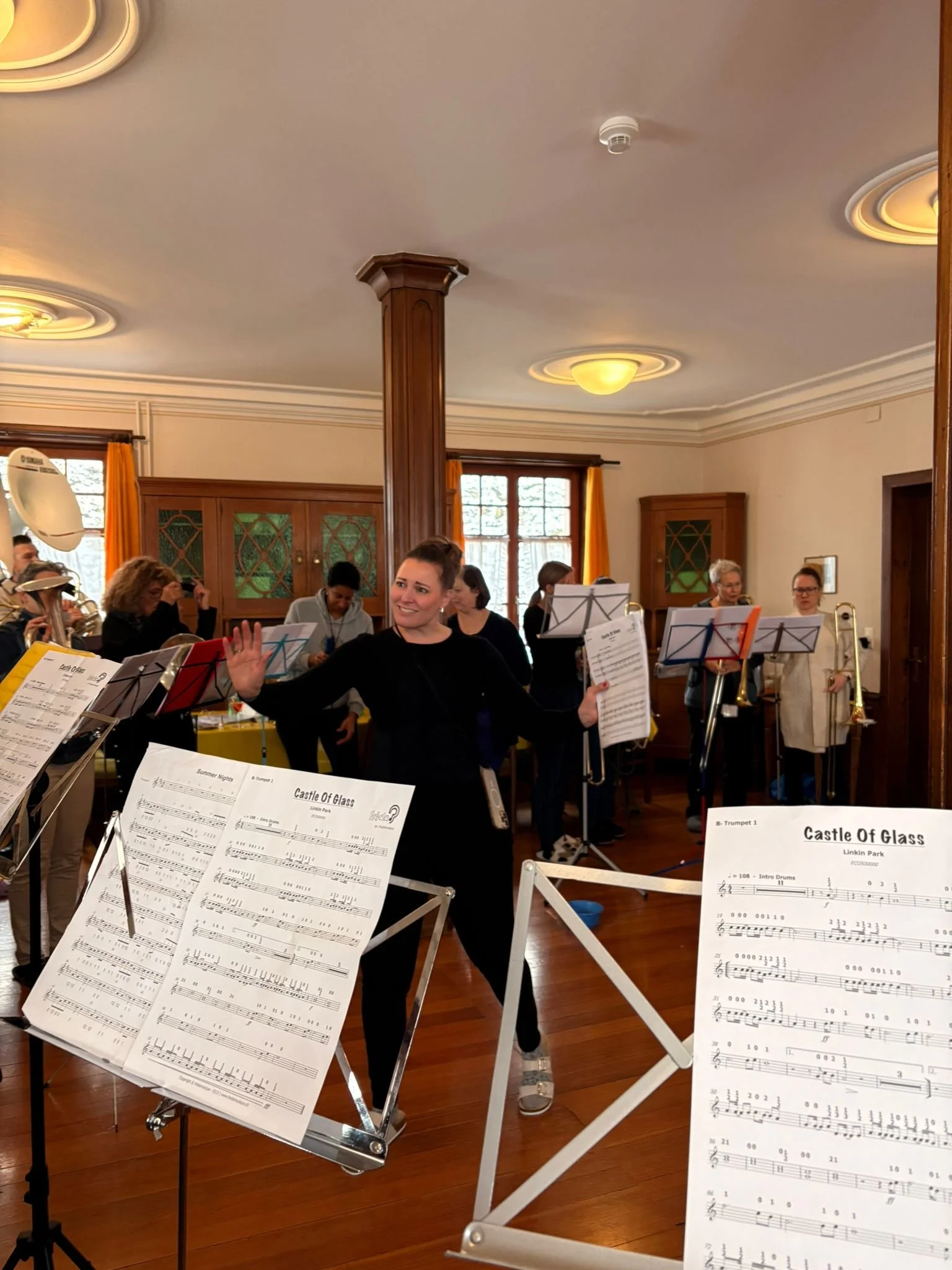 Eine Gruppe von Musikern, darunter die Dirigentin, bereitet sich auf eine Probe oder ein Konzert vor. Es befinden sich Notenblätter auf Notenständern in der Nähe.