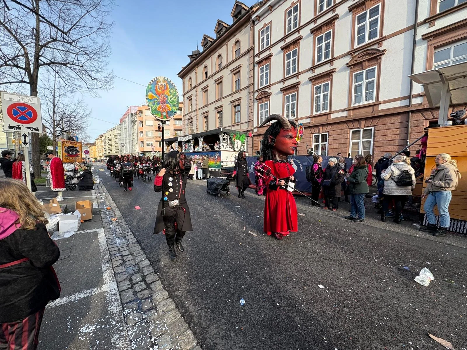 Karnevals- oder Faschingsumzug mit verkleideten Menschen, große Puppe mit Dämonenmaske, Straßenumgebung mit Gebäuden und Zuschauern.