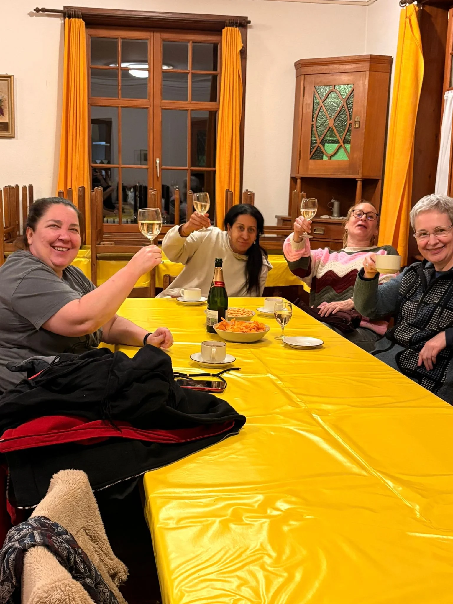 Vier Frauen feiern zusammen bei Tisch mit Sektgläsern, da vor ihnen auf einem großen gelben Tisch Getränke, Snacks und eine Flasche stehen, in einem gemütlichen Raum mit gelben Vorhängen und Holzmöbeln.