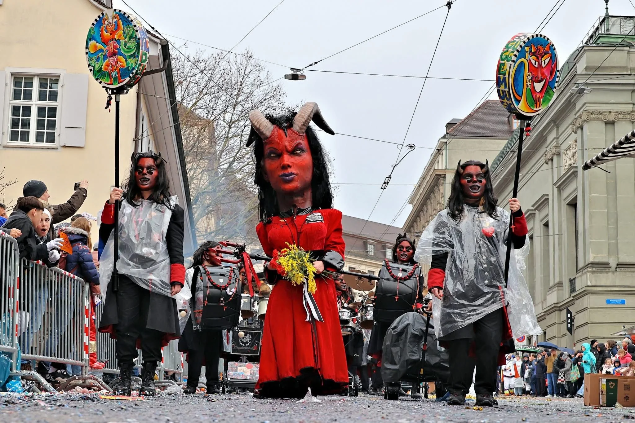 Menschen in Karnevalskostümen, darunter eine mit Teufelmaske und roten Kleid, die in einem Fasnachtsumzug marschieren, umgeben von Zuschauern.