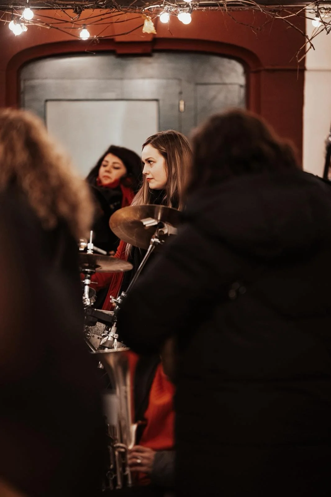 Mehrere Menschen, darunter eine junge Frau mit langen braunen Haaren, sitzen in einer entspannten Atmosphäre, wahrscheinlich bei einem Musik- oder Karaoke-Abend, in einem cozy Raum mit dunklem Holz und Lichterketten.