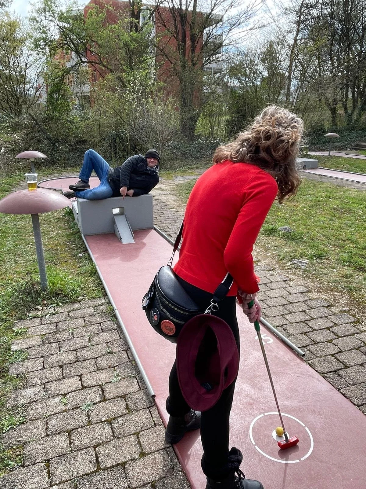 Frau spielt Mini-Golf auf einem kleinen, roten Abschlag, während ein Mann entspannt auf einer Skateboard-ähnlichen Plattform liegt. Der Hintergrund zeigt Bäume und einen Gartenanschnitt.