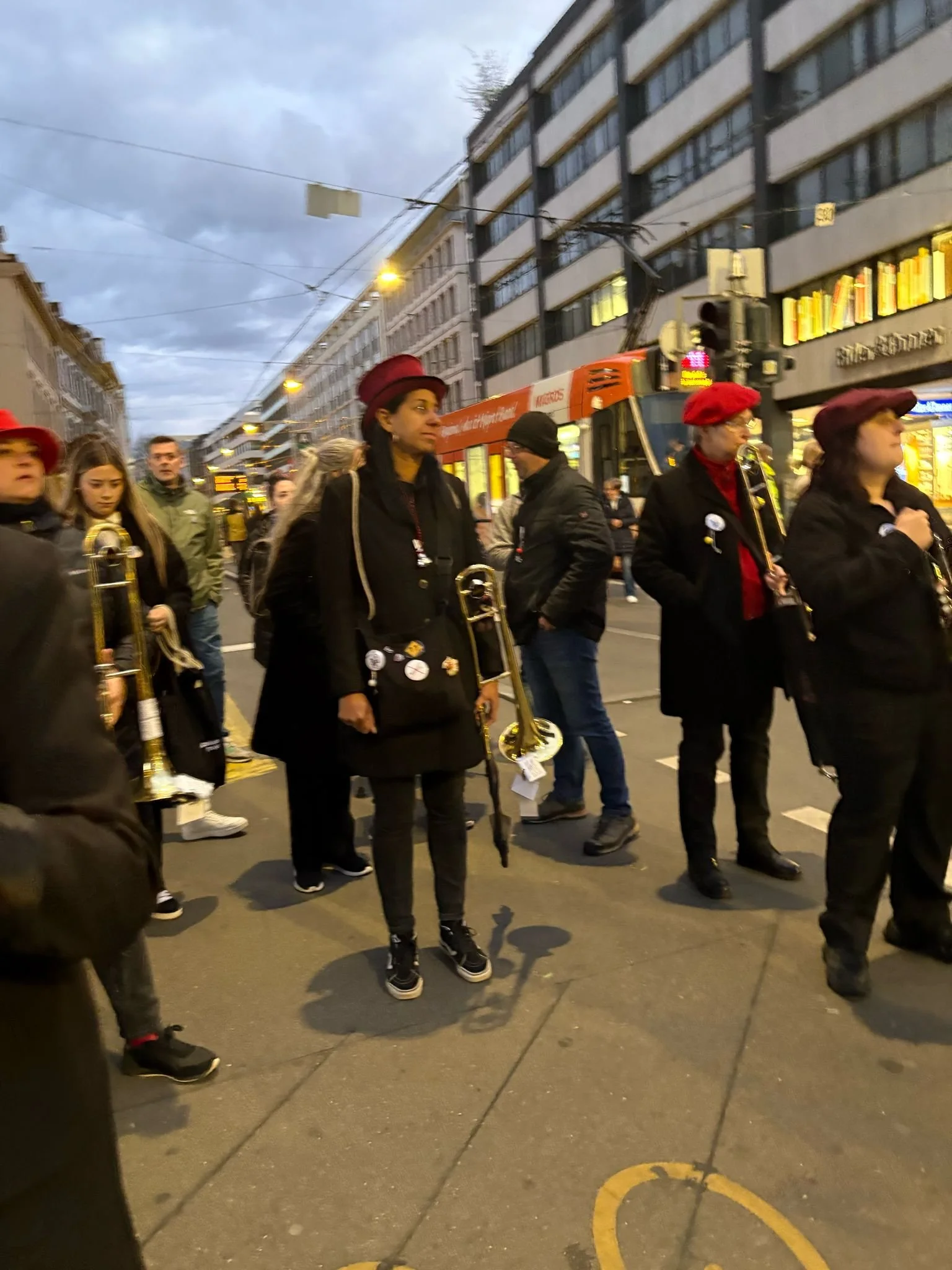 Menschen mit Musikinstrumenten, vermutlich bei einem Umzug oder Auftritt, auf einer Straßenkreuzung in einer Stadt am Abend.