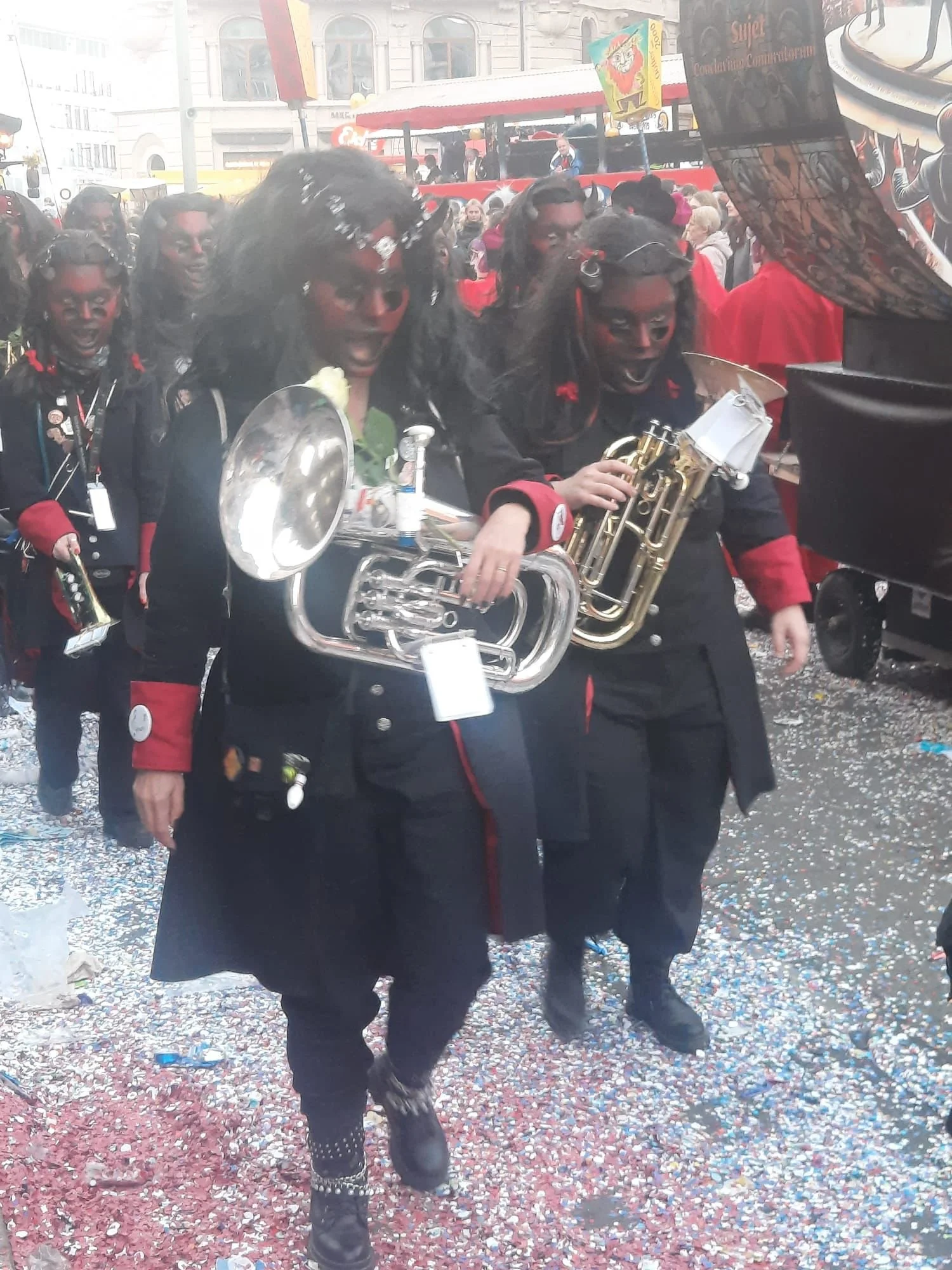 Menschen in Karnevalskostümen, musizieren auf einer Parade, schwarze Kleidung mit rotem Akzent, Konfetti auf dem Boden, festliche Stimmung.