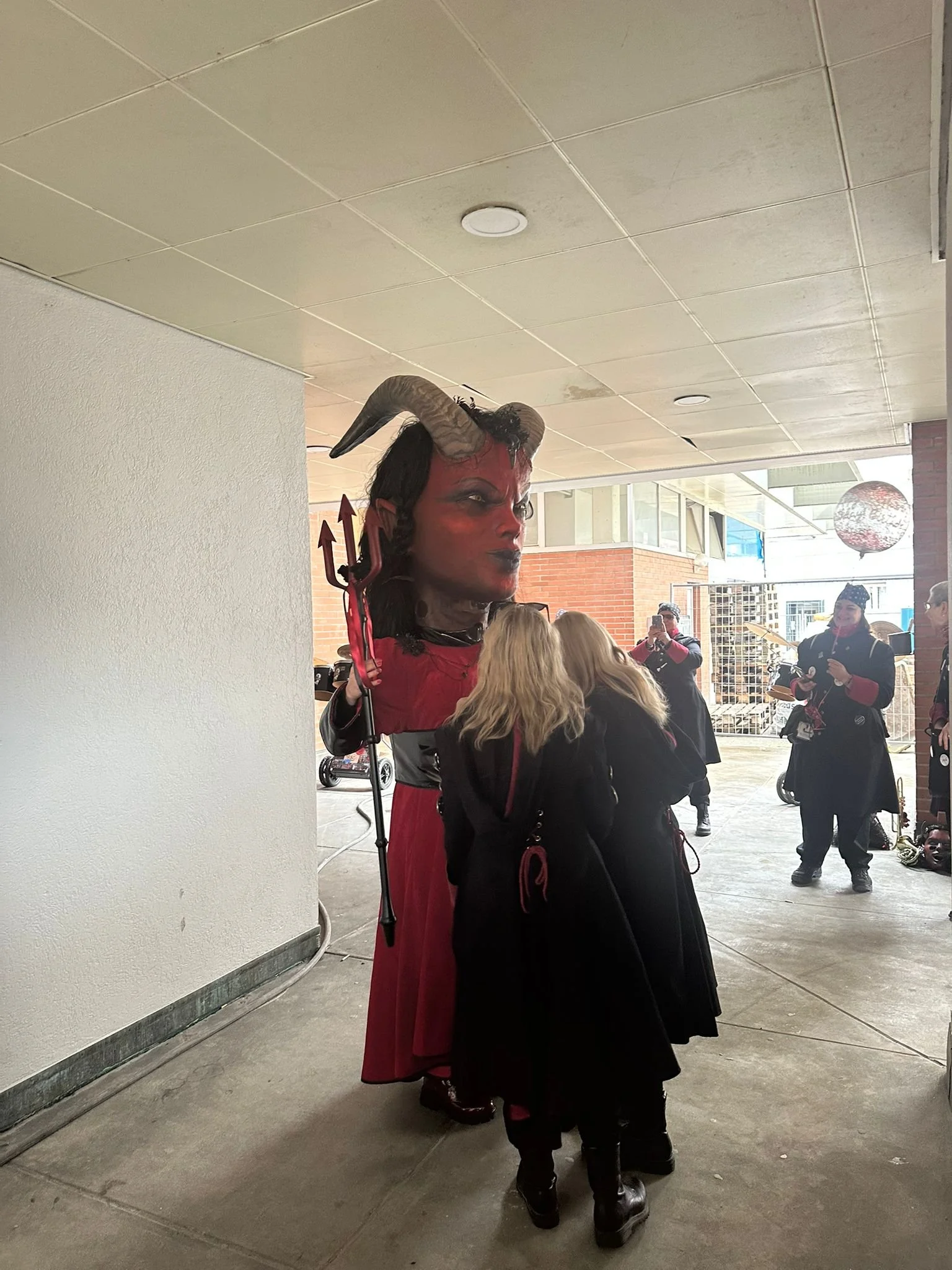 Person in Dämonenkostüm mit großen Hörnern und roter Haut, umgeben von anderen Menschen in dunkler Kleidung bei einer Feier oder Veranstaltung