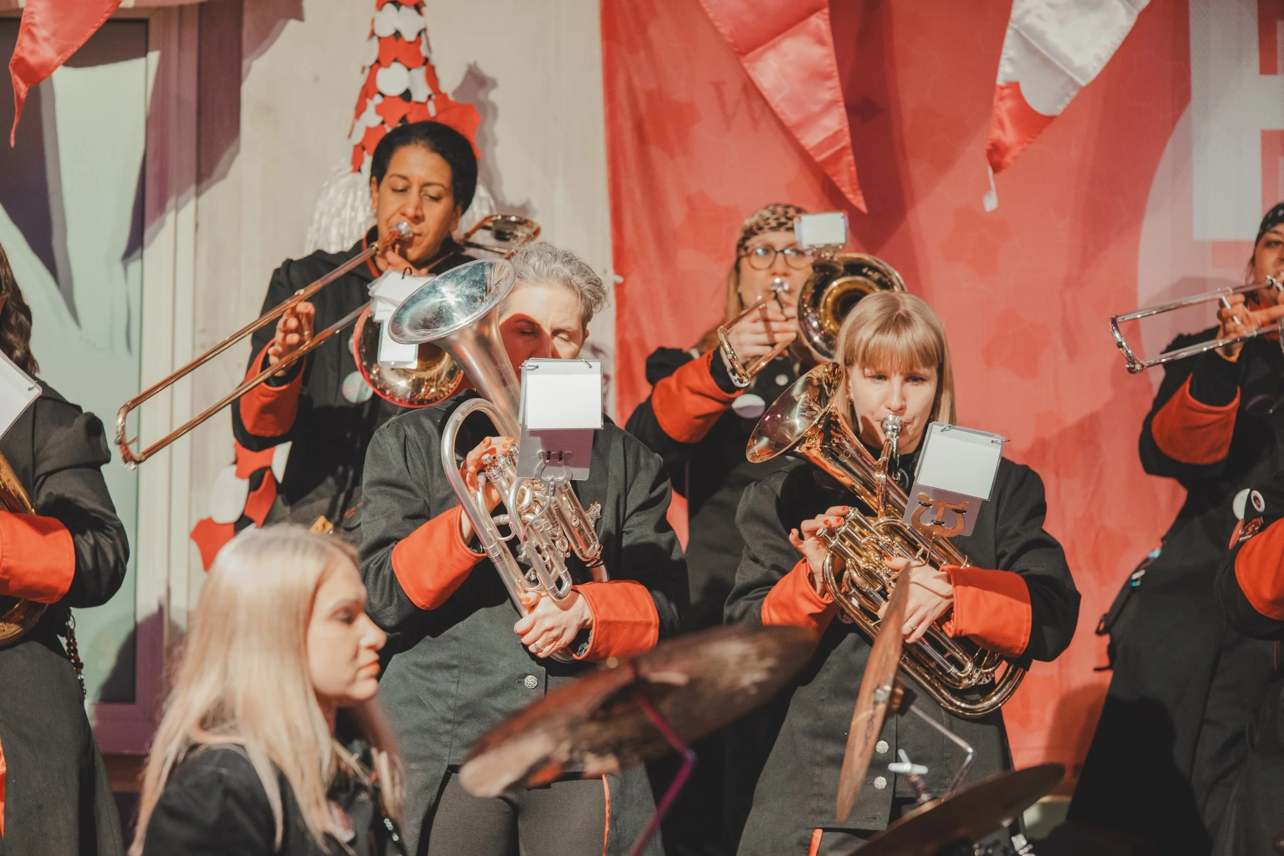 Eine Gruppe von Musikerinnen und Musikern spielt auf einer Bühne, alle tragen schwarze Kleidung mit roten Akzenten. Es sind verschiedene Blasinstrumente wie Trompeten, Flügelhörner, Posaunen und Trommeln sichtbar.