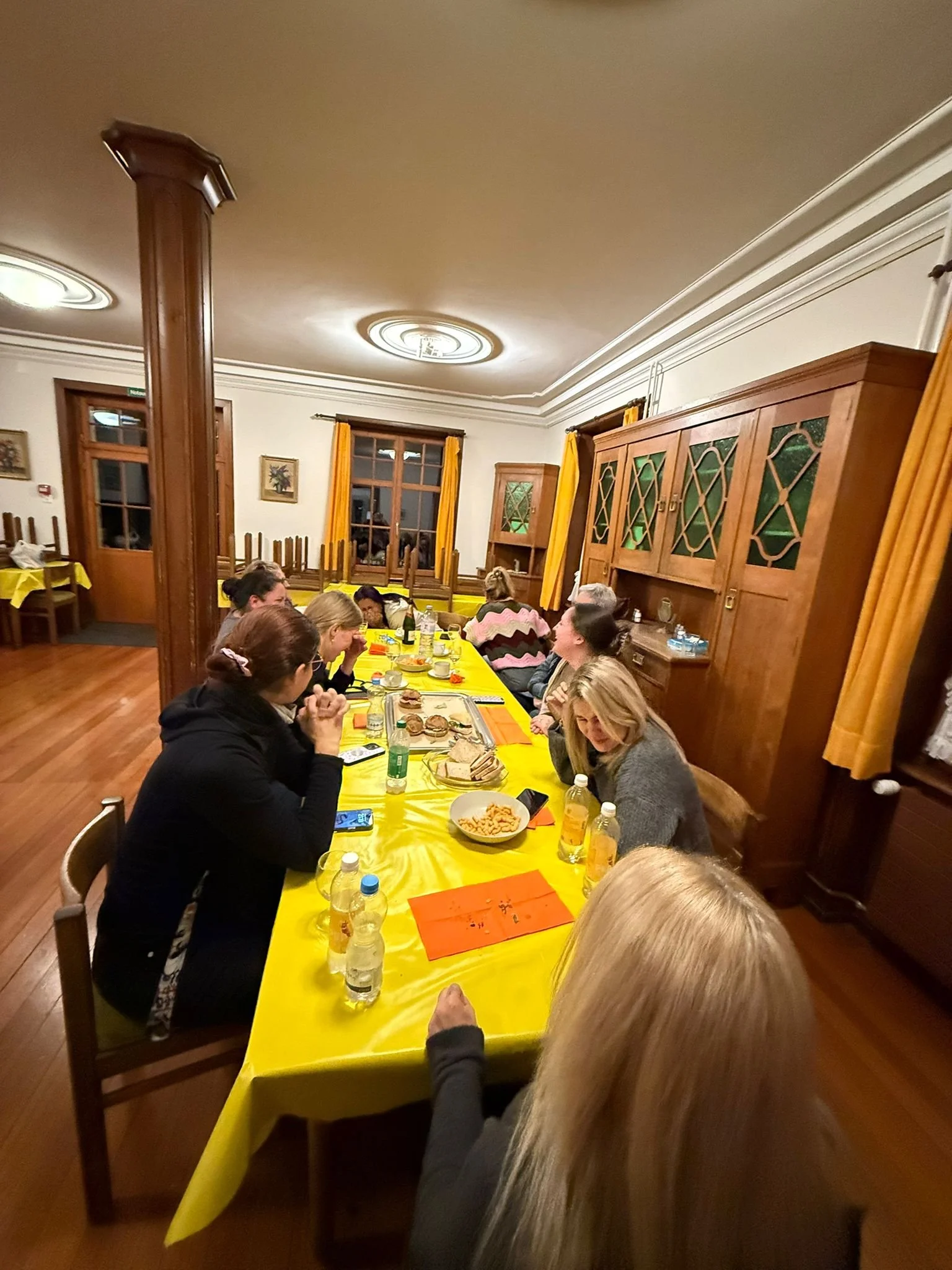 Menschen sitzen an einem langen Tisch mit gelber Tischdecke in einem festlich dekorierten Raum und essen zusammen.