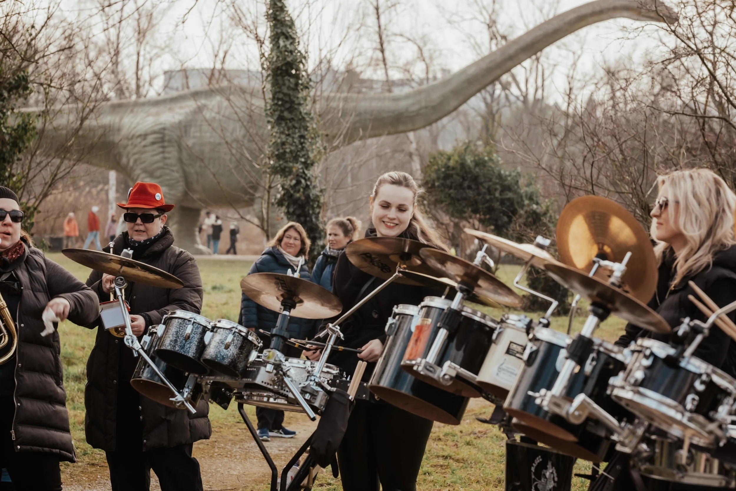 Mehrere Menschen spielen Schlagzeug im Freien, im Hintergrund ein Dinosaurier-Dino, Bäume und eine Wiese.