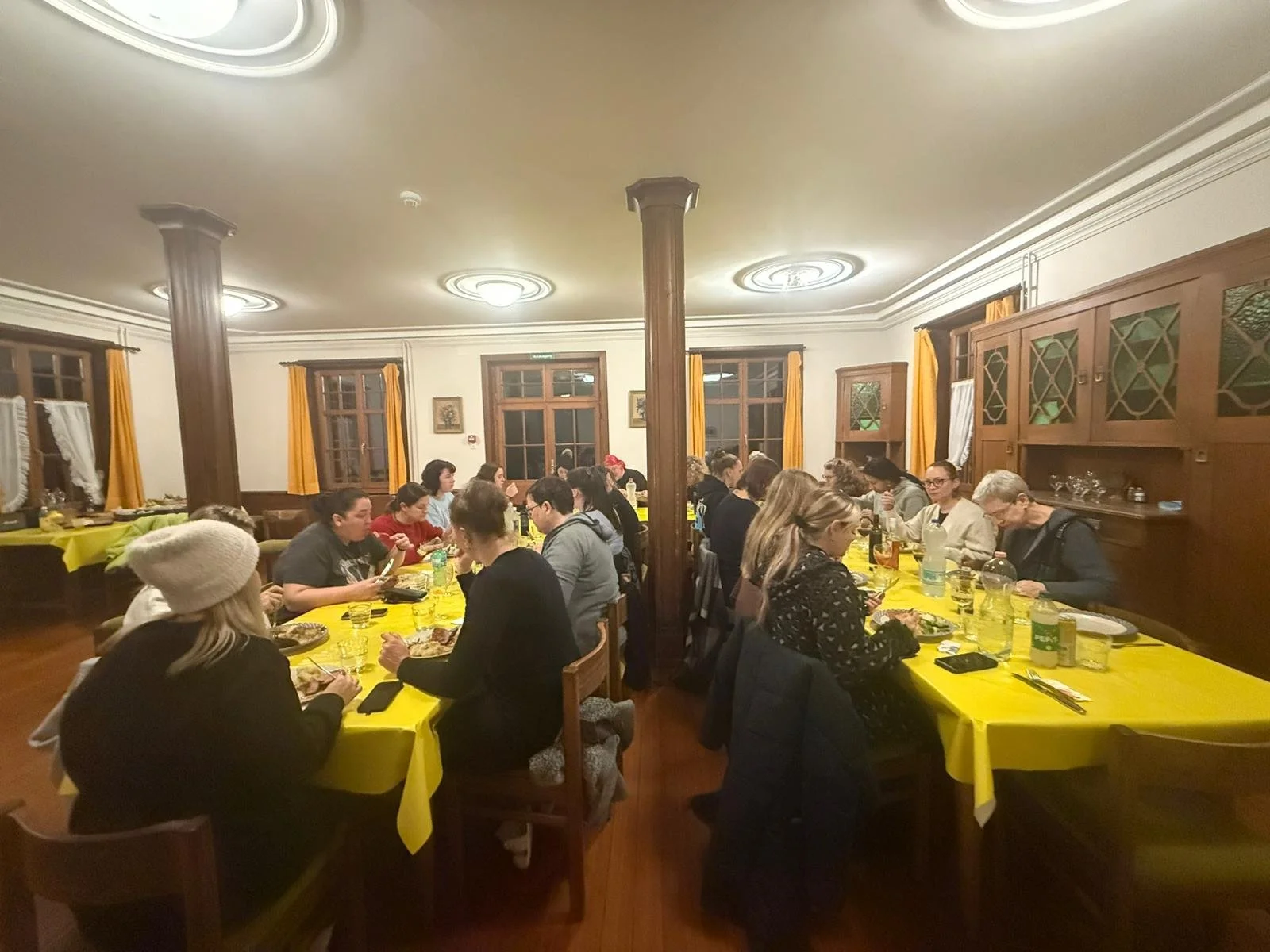Menschen sitzen an einem langen, mit gelben Tischdecken bedeckten Esstisch in einem rustikalen Raum mit Holzvertäfelung und Fenstern mit gelben Vorhängen, beim Essen.