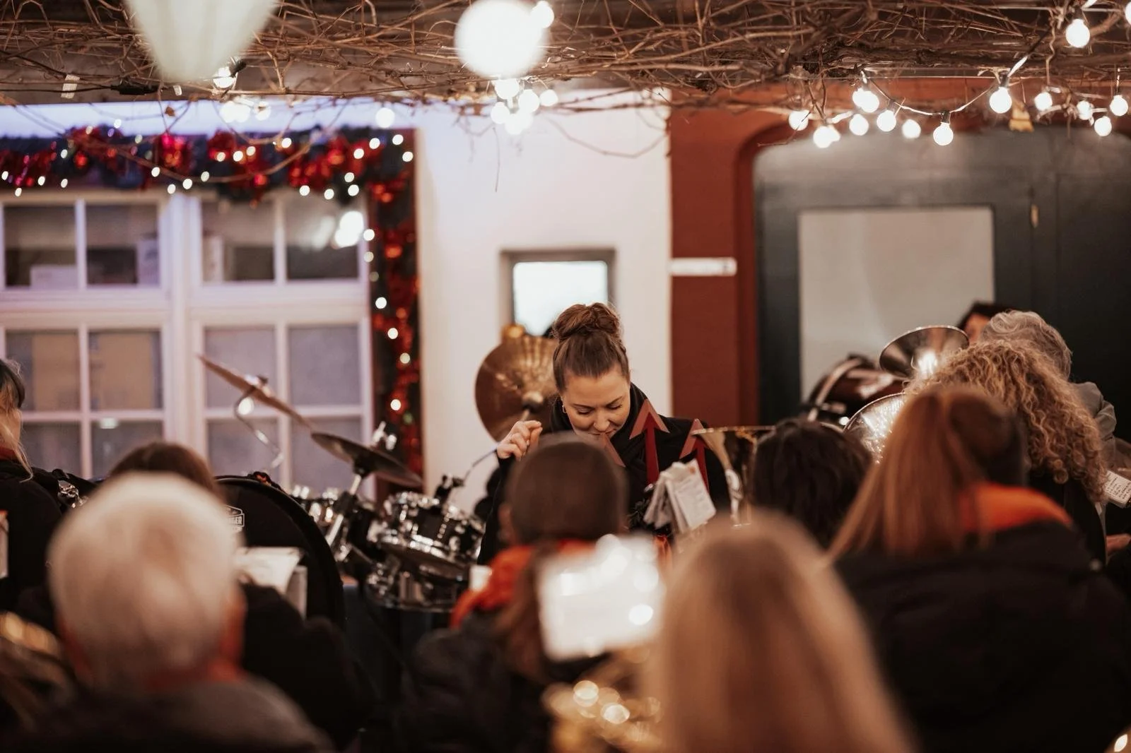 Eine Gruppe von Menschen, die in einem festlich dekorierten Raum ein Konzert mit einer Blaskapelle oder einem Musikensemble hören. Im Vordergrund sind die Köpfe der Zuhörer zu sehen, im Hintergrund eine Musikerin, die an ihrem Instrument spielt, mit 