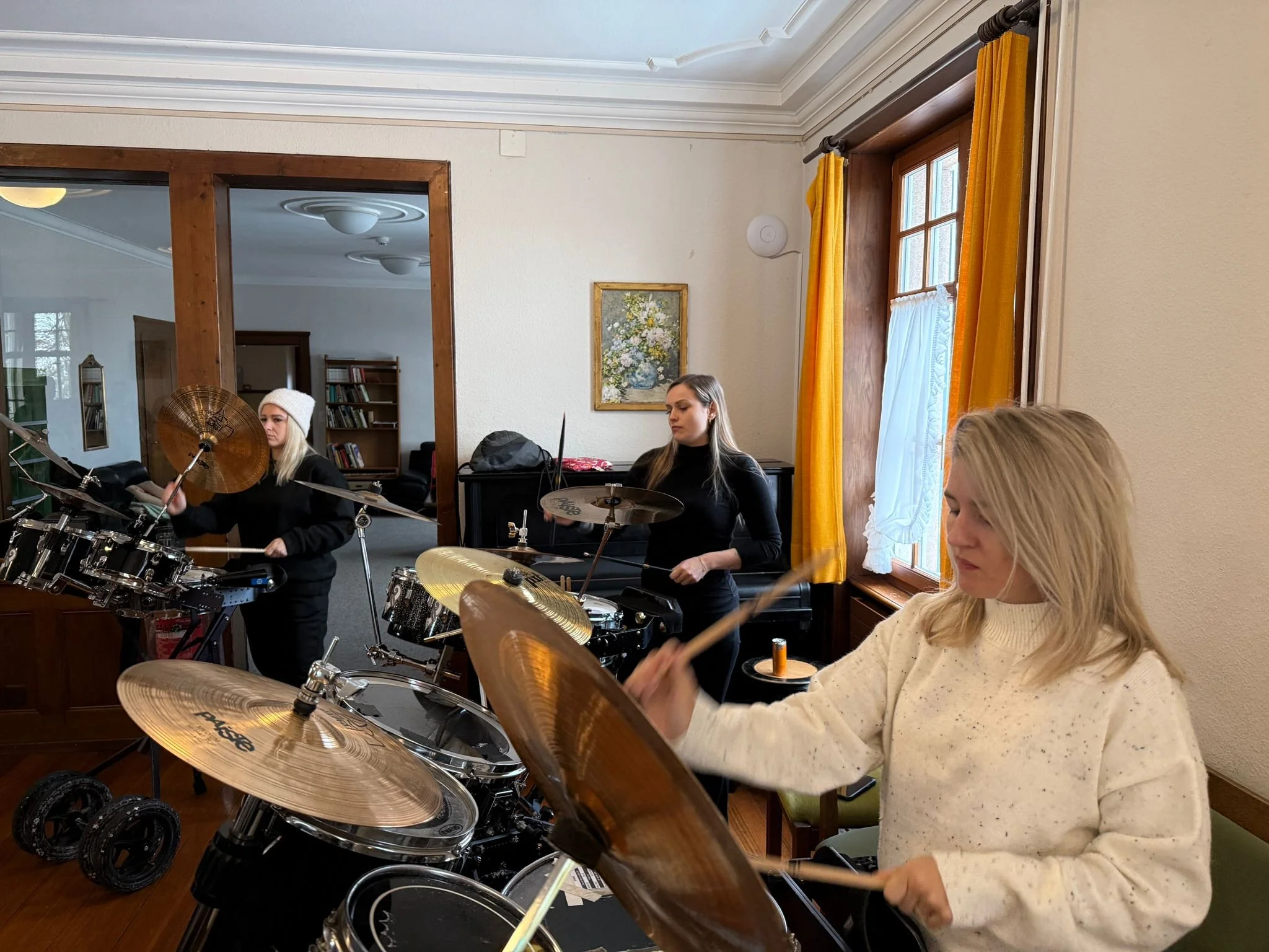 Drei junge Frauen spielen auf einem Schlagzeug, während die andere auf einer Klavierseite sitzt, im Innenraum eines Hauses mit holzvertäfelten Fenstern und gelben Vorhängen.