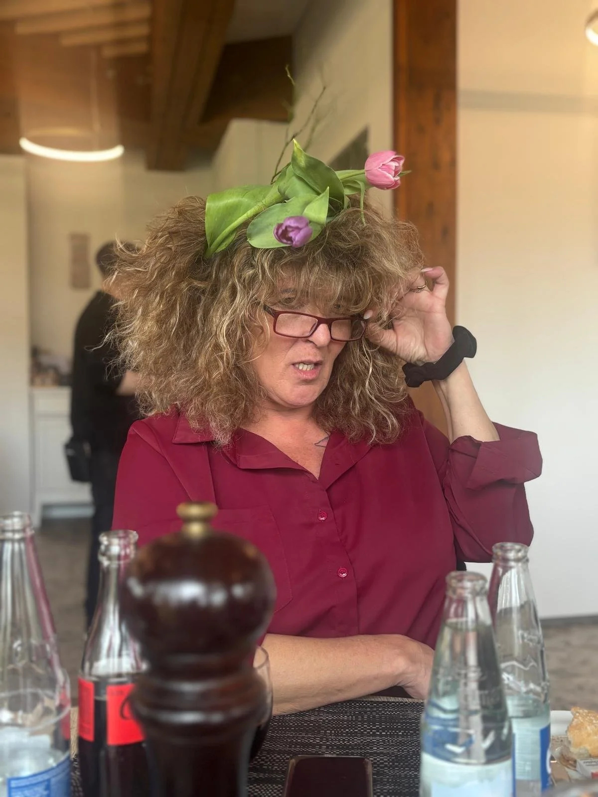 Eine Frau mit lockigem, blondem Haar, Brille und rotem Hemd sitzt an einem Tisch in einem Restaurant. Sie trägt einen Blumenkranz aus Tulpen auf dem Kopf.