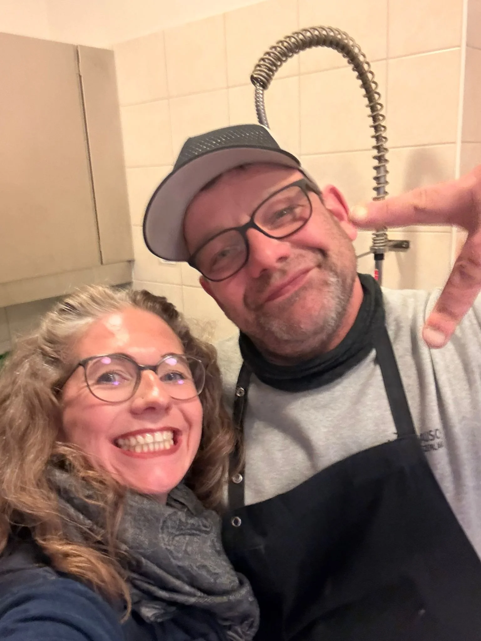 Zwei fröhliche Menschen, ein Mann und eine Frau, nehmen ein Selfie in einer Küche auf, beide tragen Brillen. Der Mann trägt eine Mütze und ein T-Shirt, die Frau trägt einen Schal.