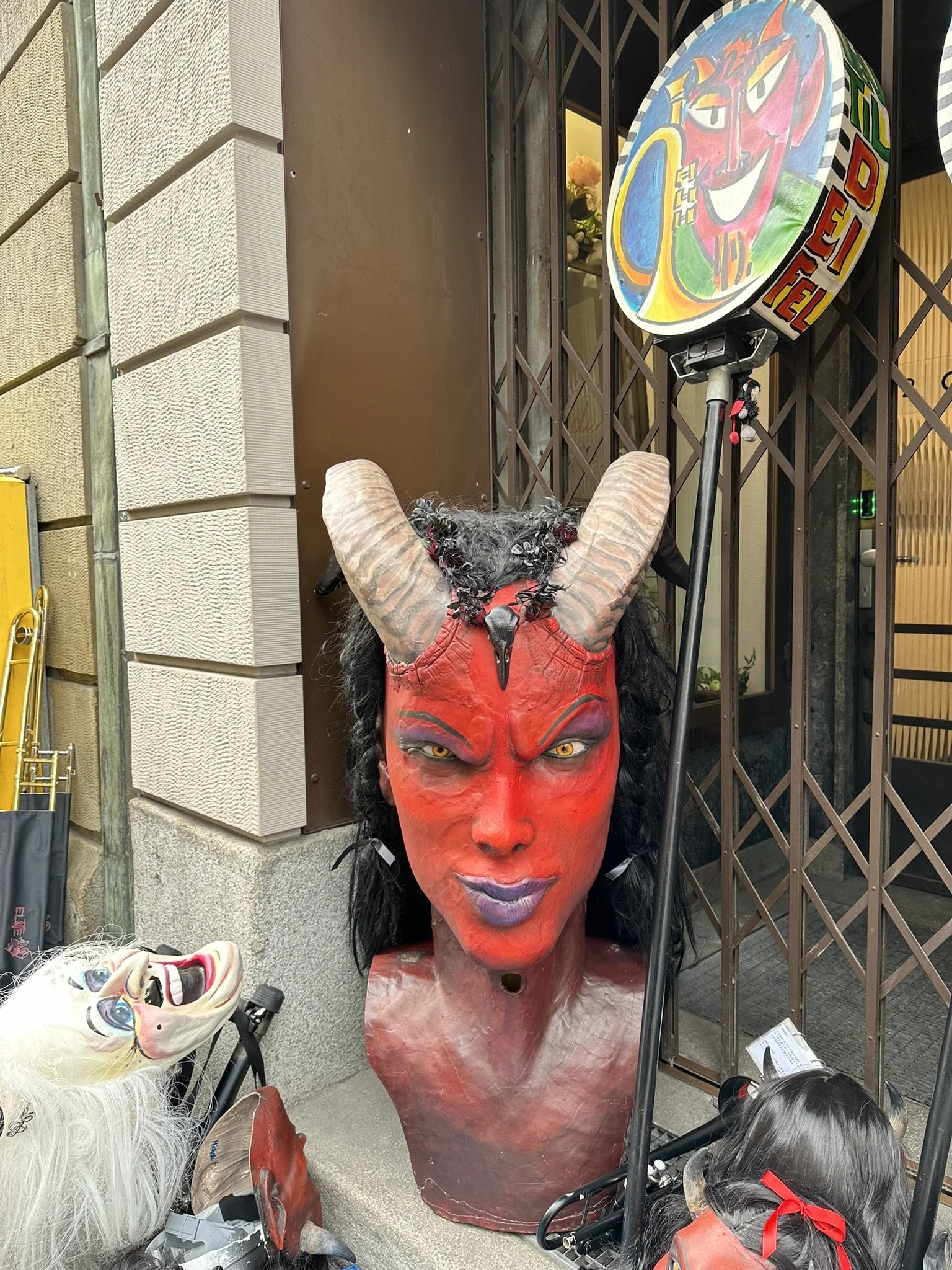 Dekorative Teufelsmaske mit roten Gesicht, schwarzen Haaren, großen Hörnern und gelben Augen. Daneben hängen andere Masken und ein bunter Verkehrszeichen mit einem dämonischen Gesicht.