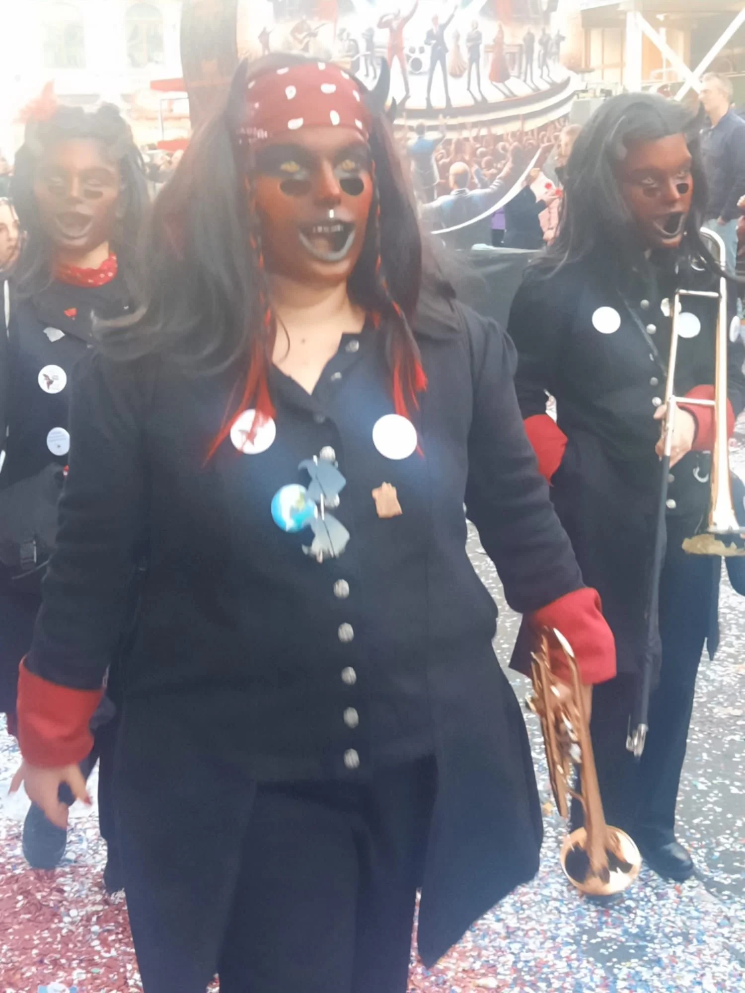 Menschen in Kostümen bei einer Feier, eine Frau im Vordergrund trägt ein schwarzes Outfit mit roten Akzenten, schwarzen Vampirzähnen, dunkler Schminke und einem roten Bandana mit weißen Punkten, im Hintergrund weitere kostümierte Personen und eine Me