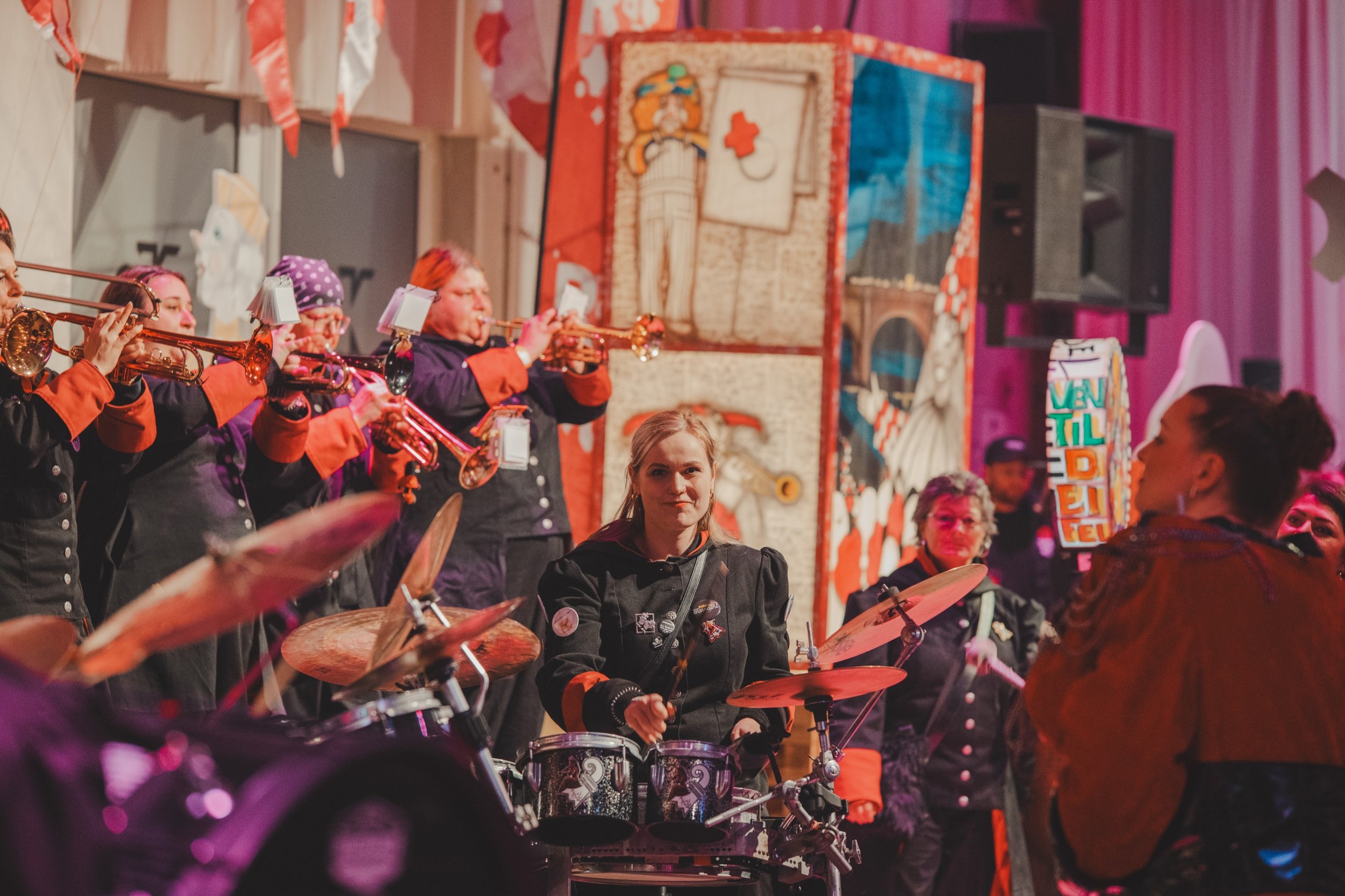 Musikerinnen bei einem Konzert, die auf einer Bühne mit buntem Hintergrund spielen, darunter Trommlerinnen, Blechbläser mit Trompeten und eine Bassistin mit einem kleineren Schlagzeug, im Hintergrund bunte Dekorationen und ein Plakat.