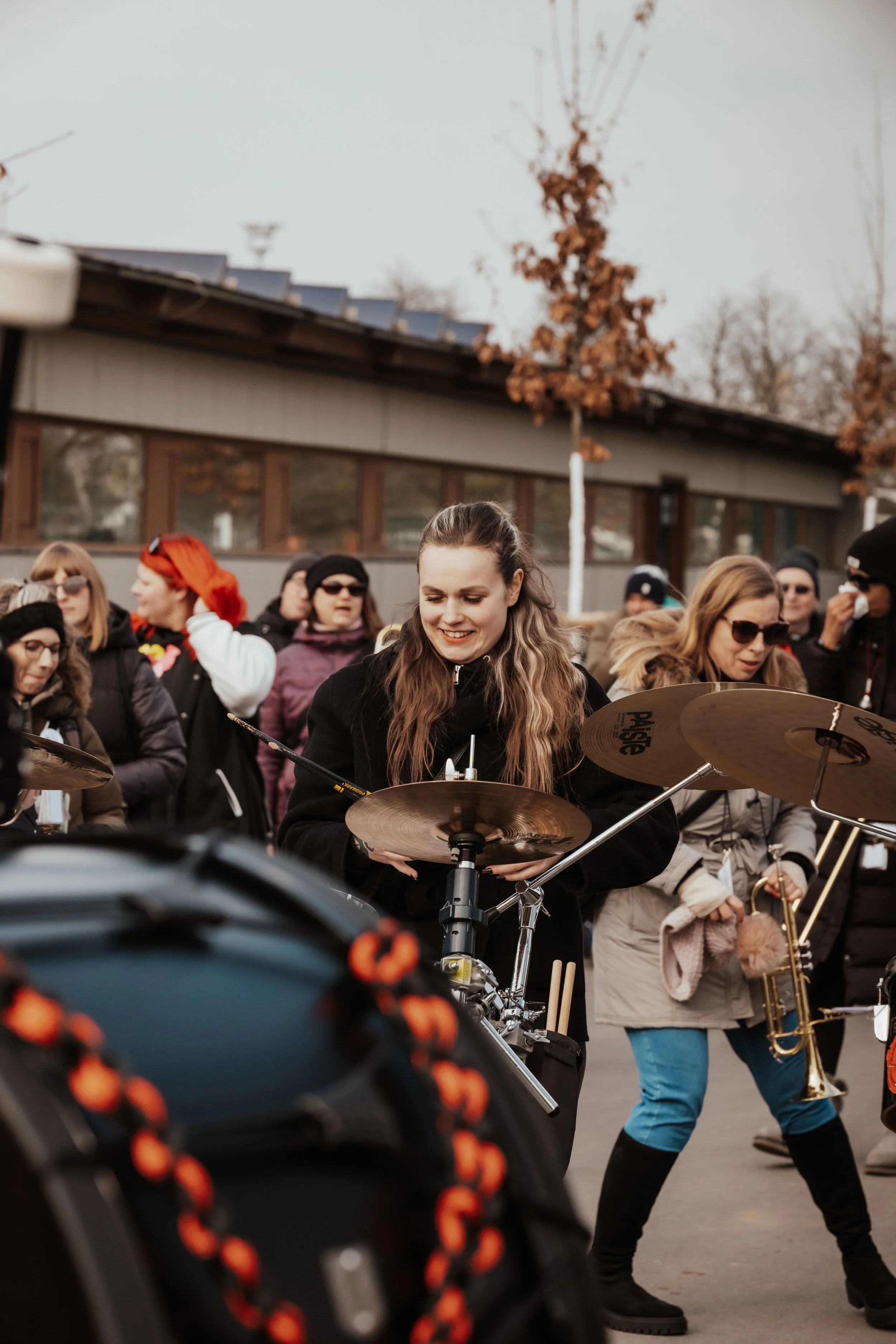 Menschen spielen Musik im Freien, darunter Schlagzeug, Trompete und andere Instrumente, bei bewölktem Himmel.