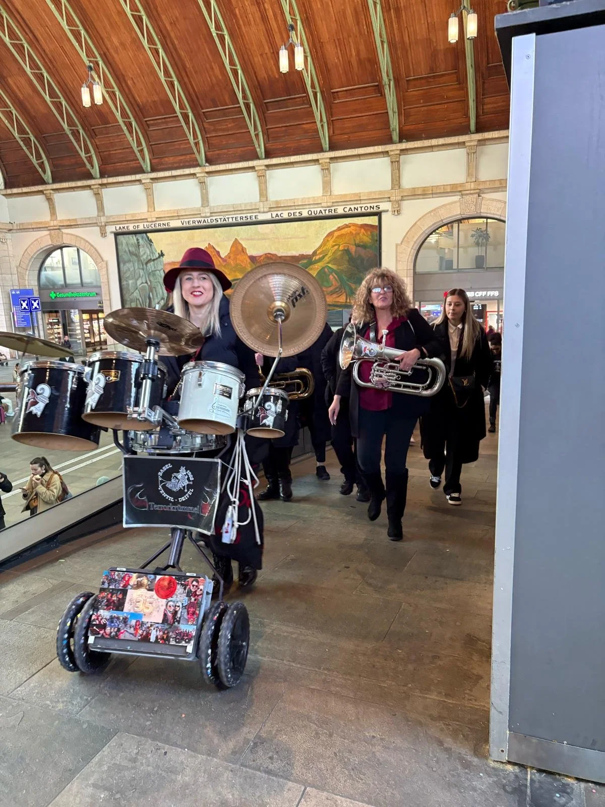 Eine Gruppe von Straßenmusikern mit Instrumenten, die durch einen Bahnhof gehen. Im Hintergrund ist eine große Wandmalerei von Bergen und ein Wandbild mit verschiedenen Sprachen.