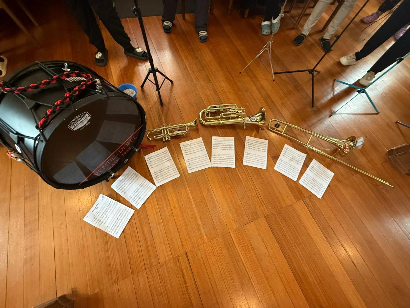 Bläserensemble mit Trommel, drei verschiedene Trompeten, Notenblätter auf dem Holzboden, und Musikständer, in einem Raum mit Holzboden.