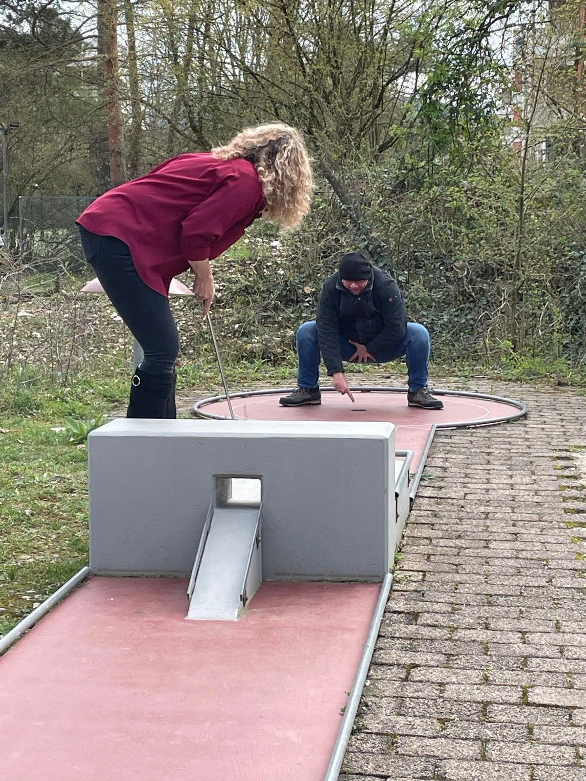 Zwei Menschen spielen Minigolf im Freien, einer steht hinter dem Loch, der andere für den nächsten Schlag, umgeben von Bäumen und einem gepflasterten Weg.