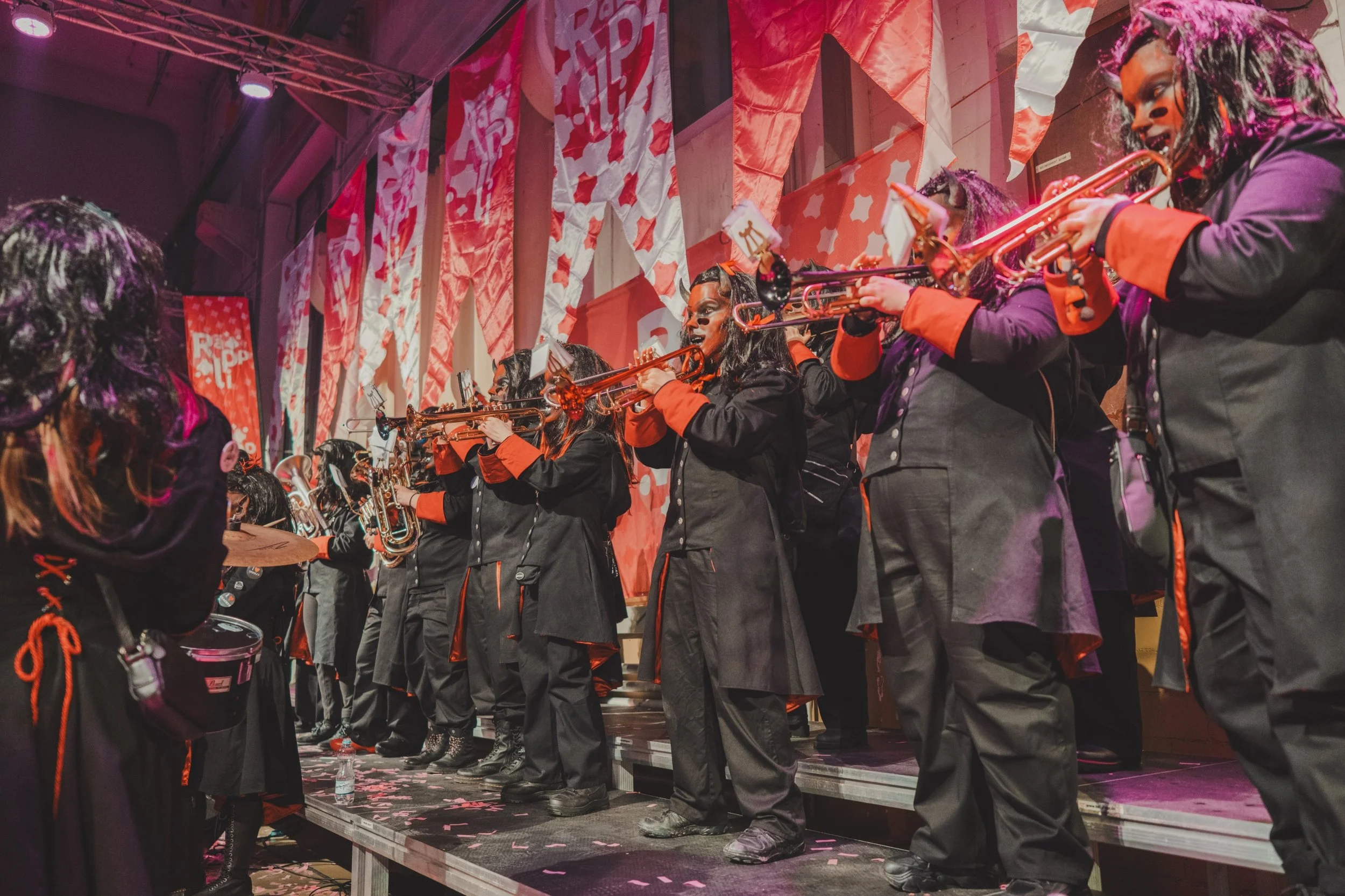 Gruppe von Musikern, die auf einer Bühne Töne mit Trompeten und einem Schlagzeug spielen, während sie in schwarzen Uniformen mit roten Akzenten gekleidet sind, im Hintergrund rote sowie weiße Bühnenverkleidungen und Dekorationen.